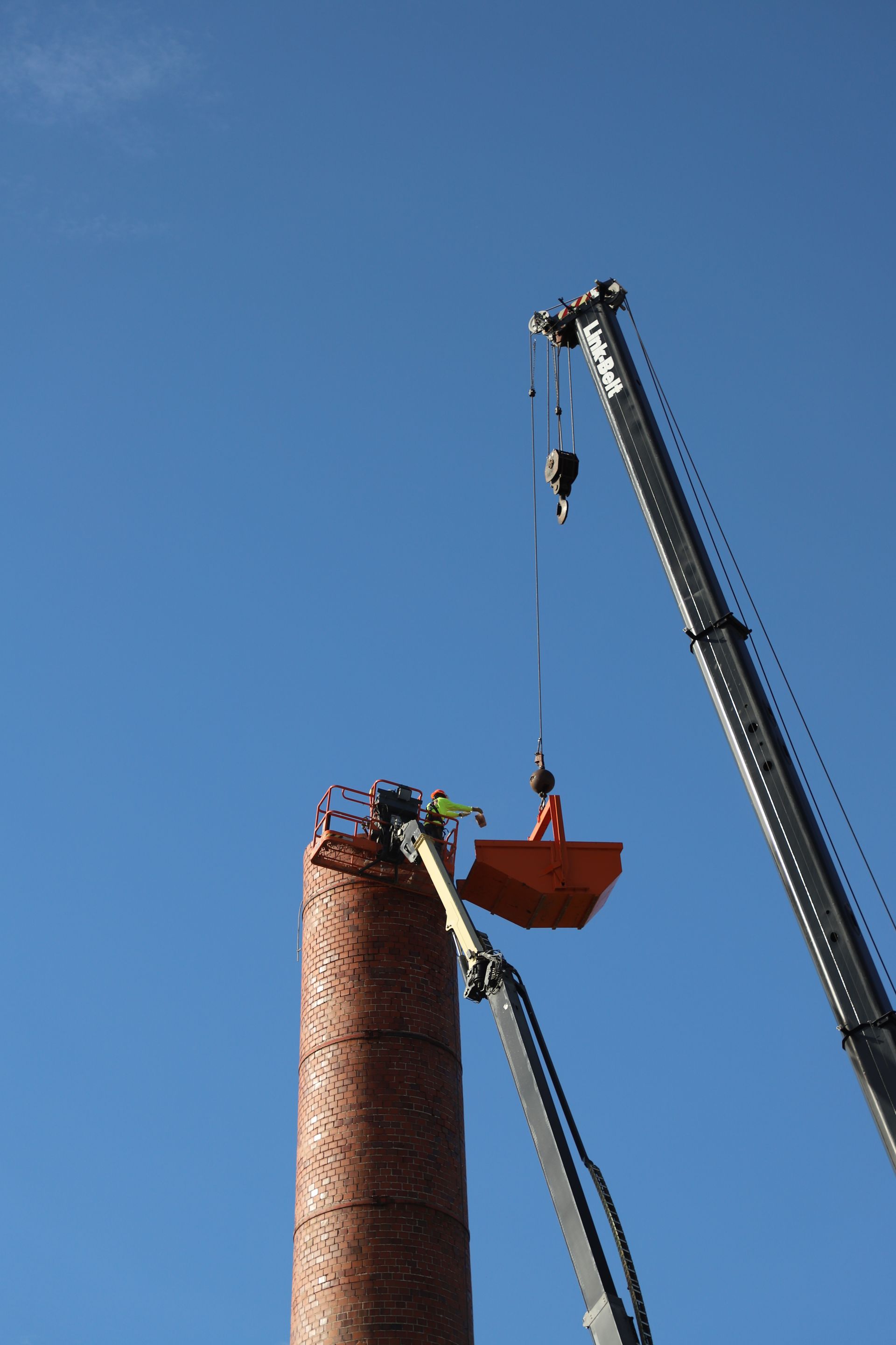 CL Construction crew performing hand demolition on a tall industrial smokestack at the Lincoln Regional Center site in Lincoln, Nebraska. Workers use manual tools and safety gear to carefully dismantle sections of the 143-foot chimney structure amid rubble and heavy equipment on the ground