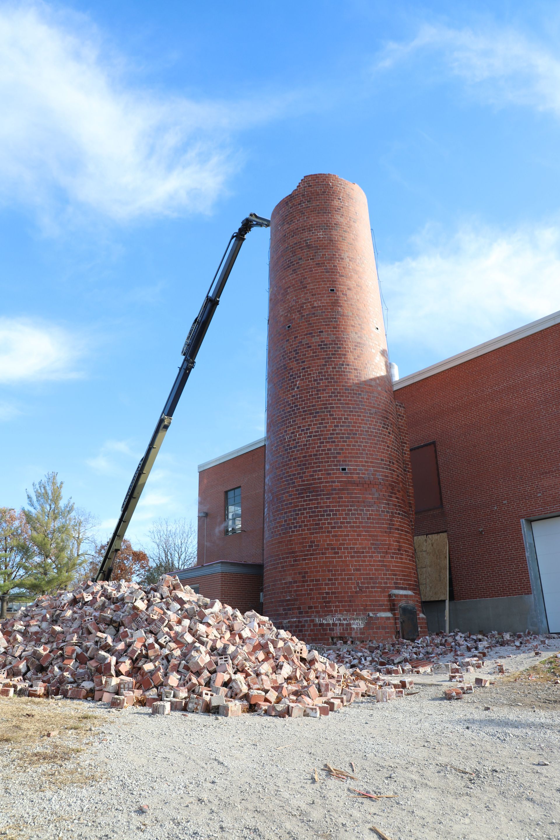 CL Construction crew performing hand demolition on a tall industrial smokestack at the Lincoln Regional Center site in Lincoln, Nebraska. Workers use manual tools and safety gear to carefully dismantle sections of the 143-foot chimney structure amid rubble and heavy equipment on the ground