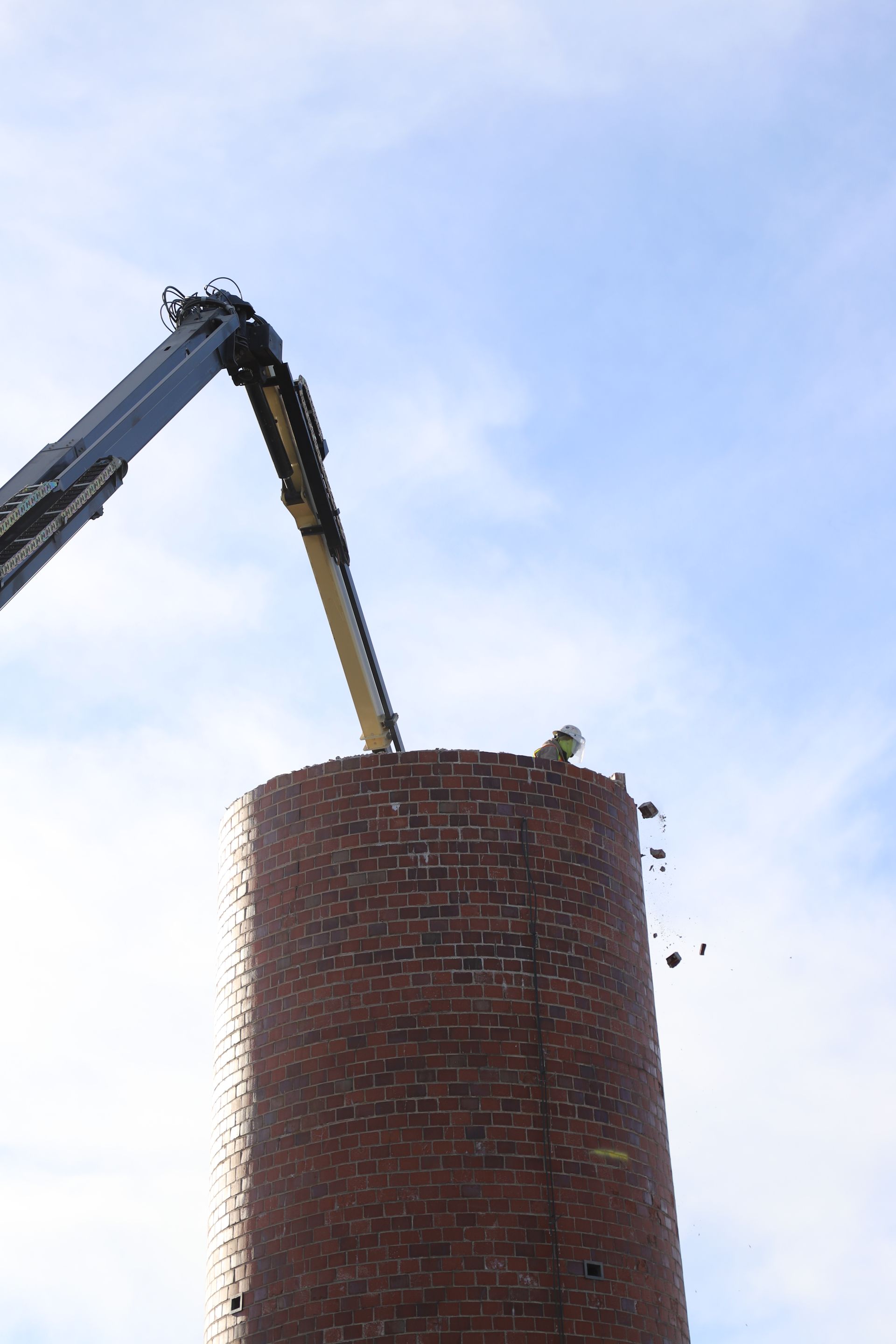 CL Construction crew performing hand demolition on a tall industrial smokestack at the Lincoln Regional Center site in Lincoln, Nebraska. Workers use manual tools and safety gear to carefully dismantle sections of the 143-foot chimney structure amid rubble and heavy equipment on the ground
