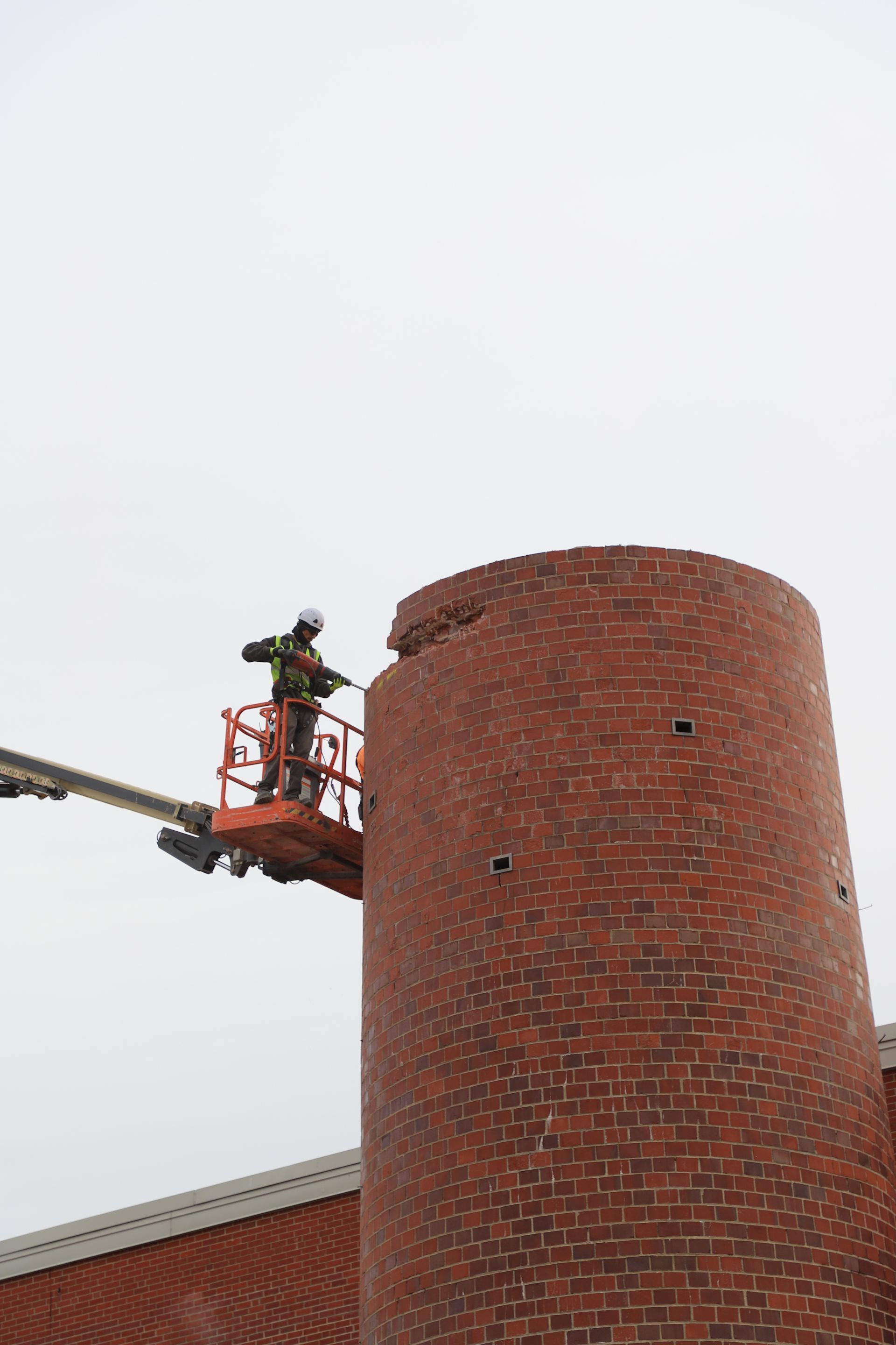 CL Construction crew performing hand demolition on a tall industrial smokestack at the Lincoln Regional Center site in Lincoln, Nebraska. Workers use manual tools and safety gear to carefully dismantle sections of the 143-foot chimney structure amid rubble and heavy equipment on the ground