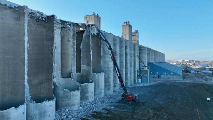 A large concrete wall is being demolished by a crane