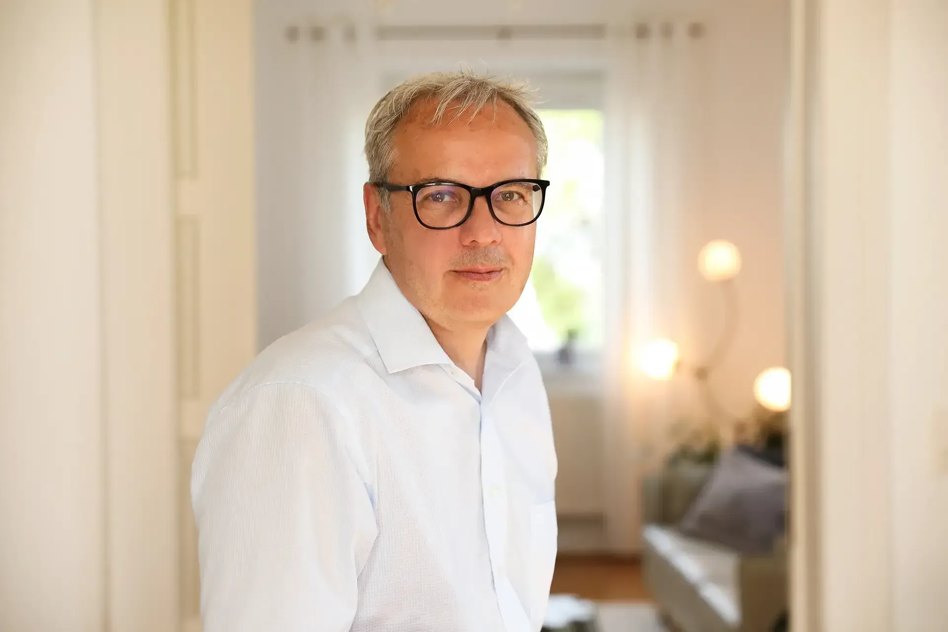 Ein Mann mit Brille und weißem Hemd steht in einem schwach beleuchteten Flur und schaut in die Kamera.