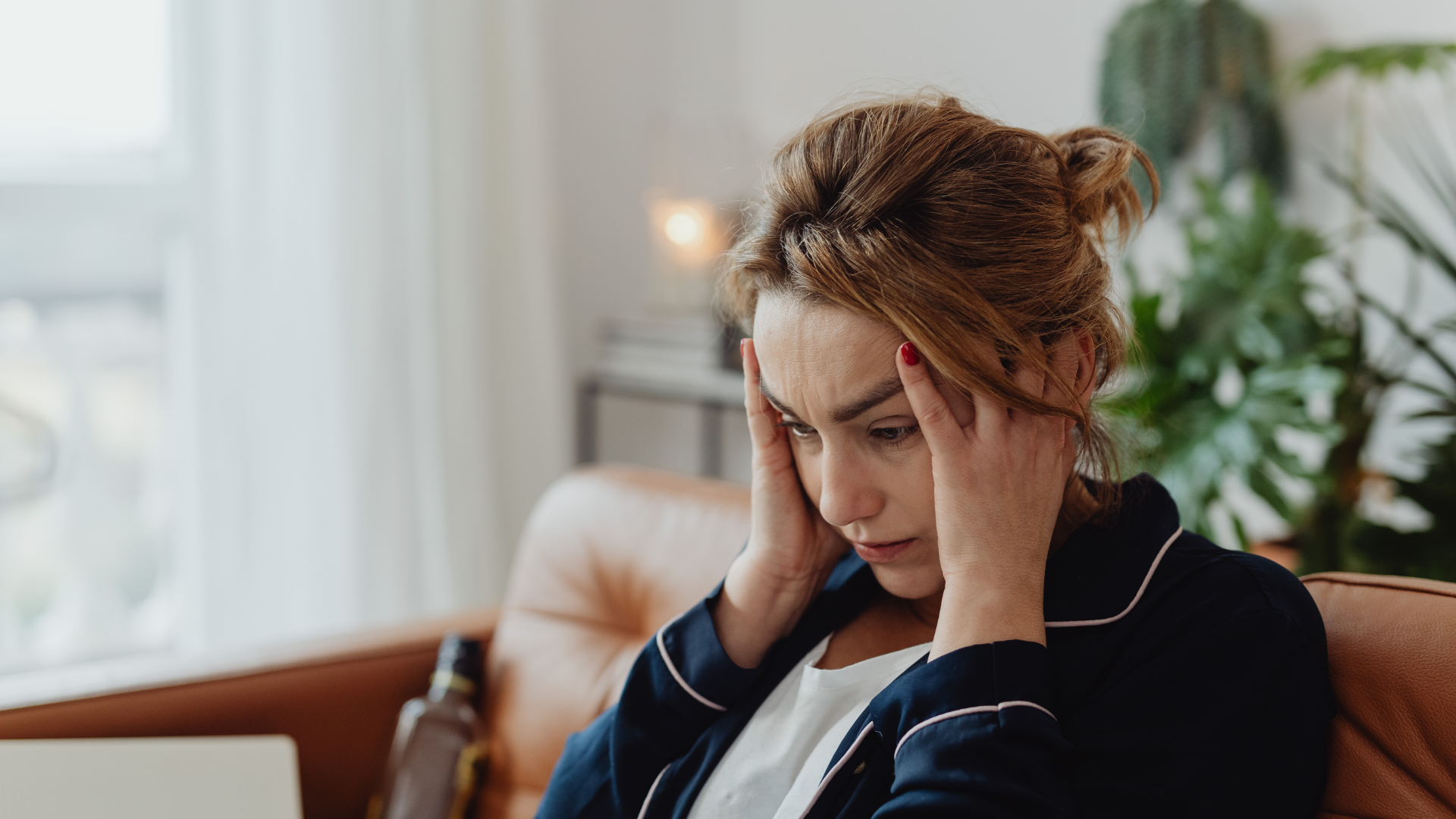 A person in pajamas sits on a brown couch with their hands on their head, appearing distressed in a home setting.