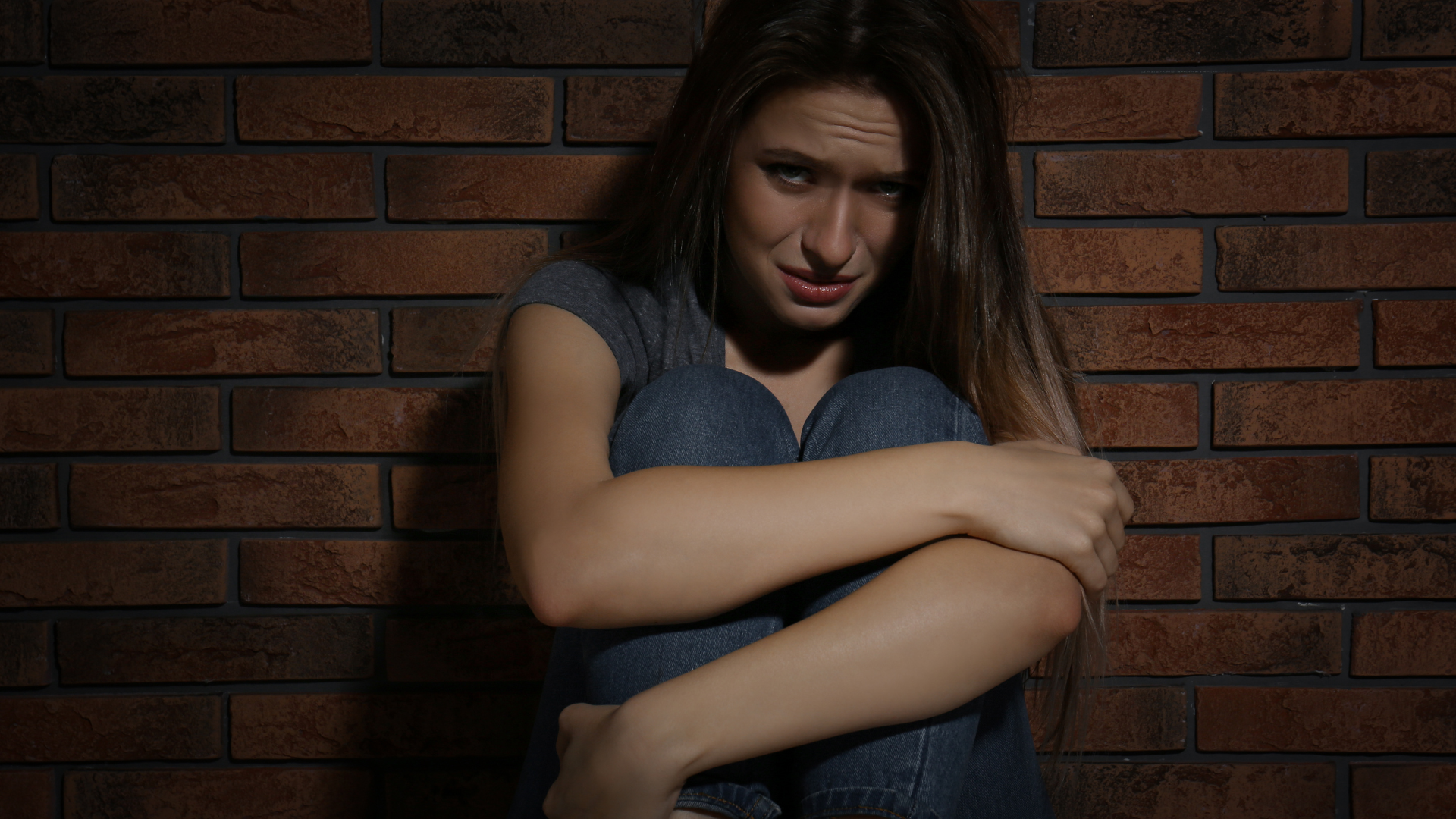 A person sitting against a brick wall, hugging their knees with a distressed expression.