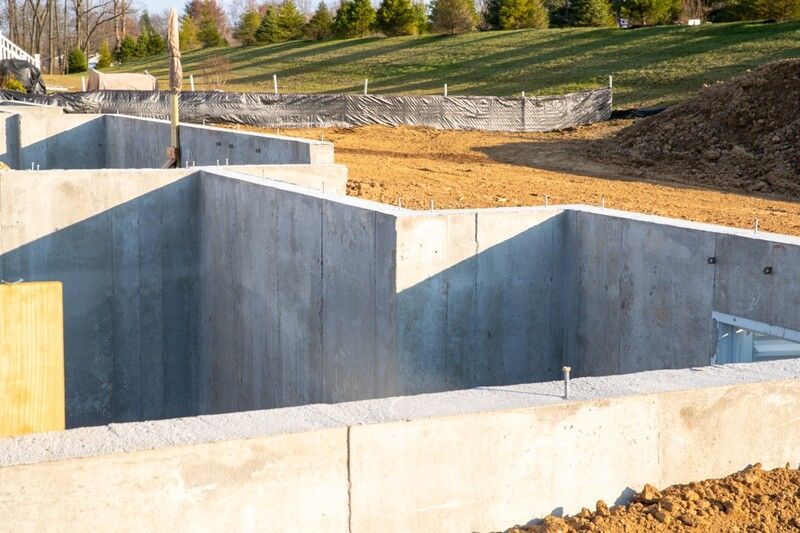Concrete foundation walls for a building under construction on a dirt lot, with grass and trees in the background