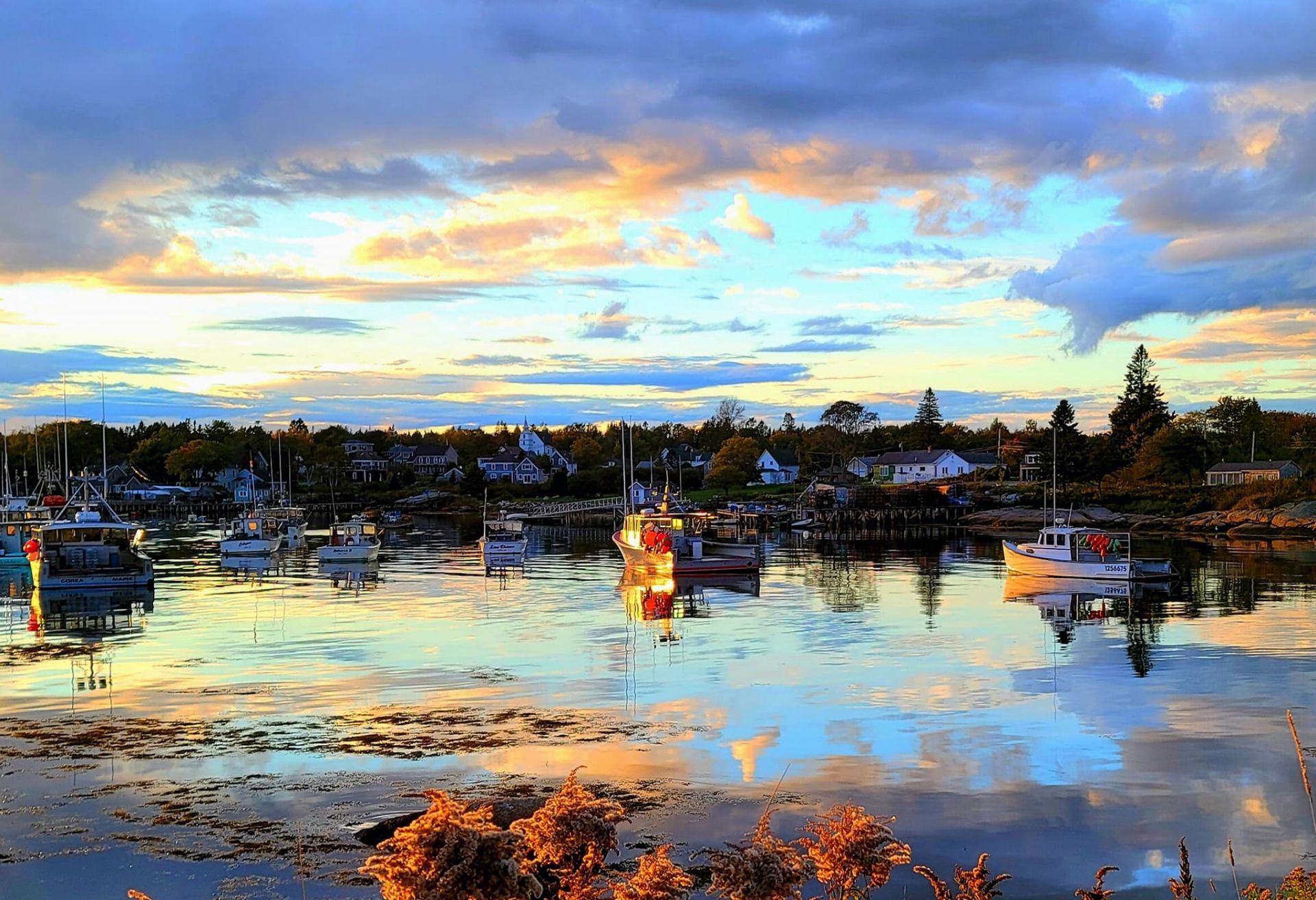 Harbor at sunset, boats docked, calm water reflects sky with gold and blue hues.