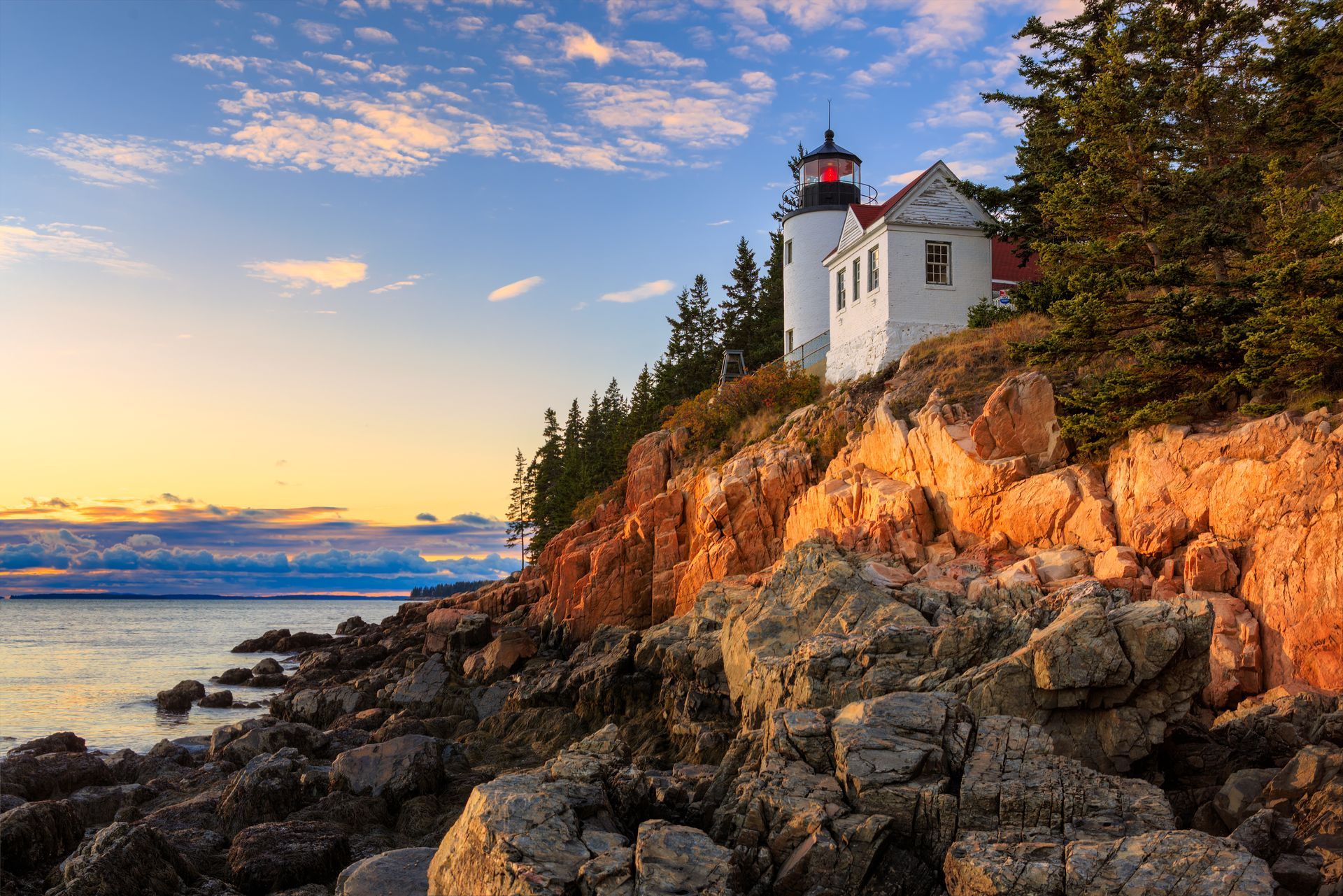 Lighthouse on a rocky cliff at sunset, overlooking the ocean with orange and blue hues.