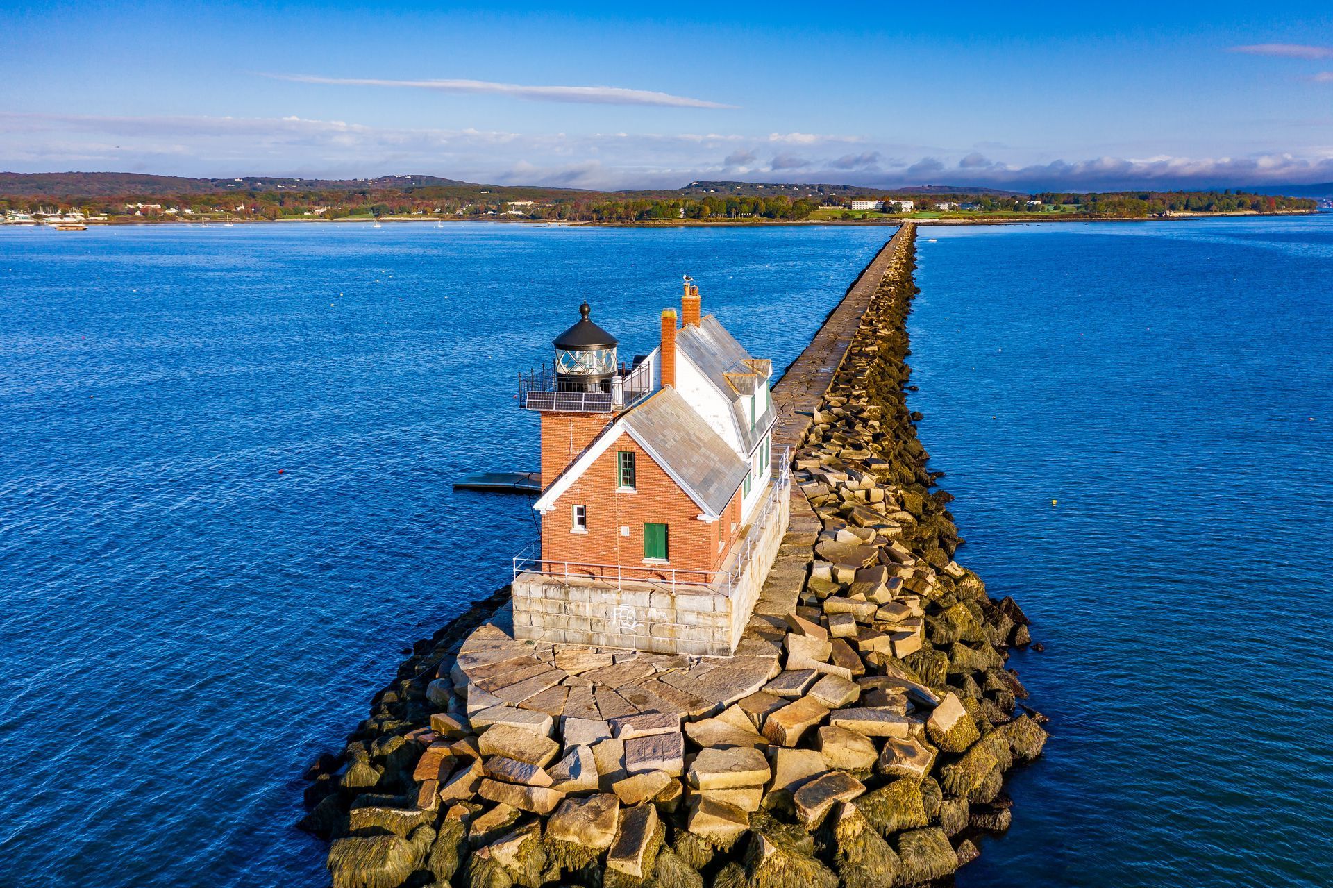 Lighthouse on rocky breakwater extends into blue water under a clear sky.