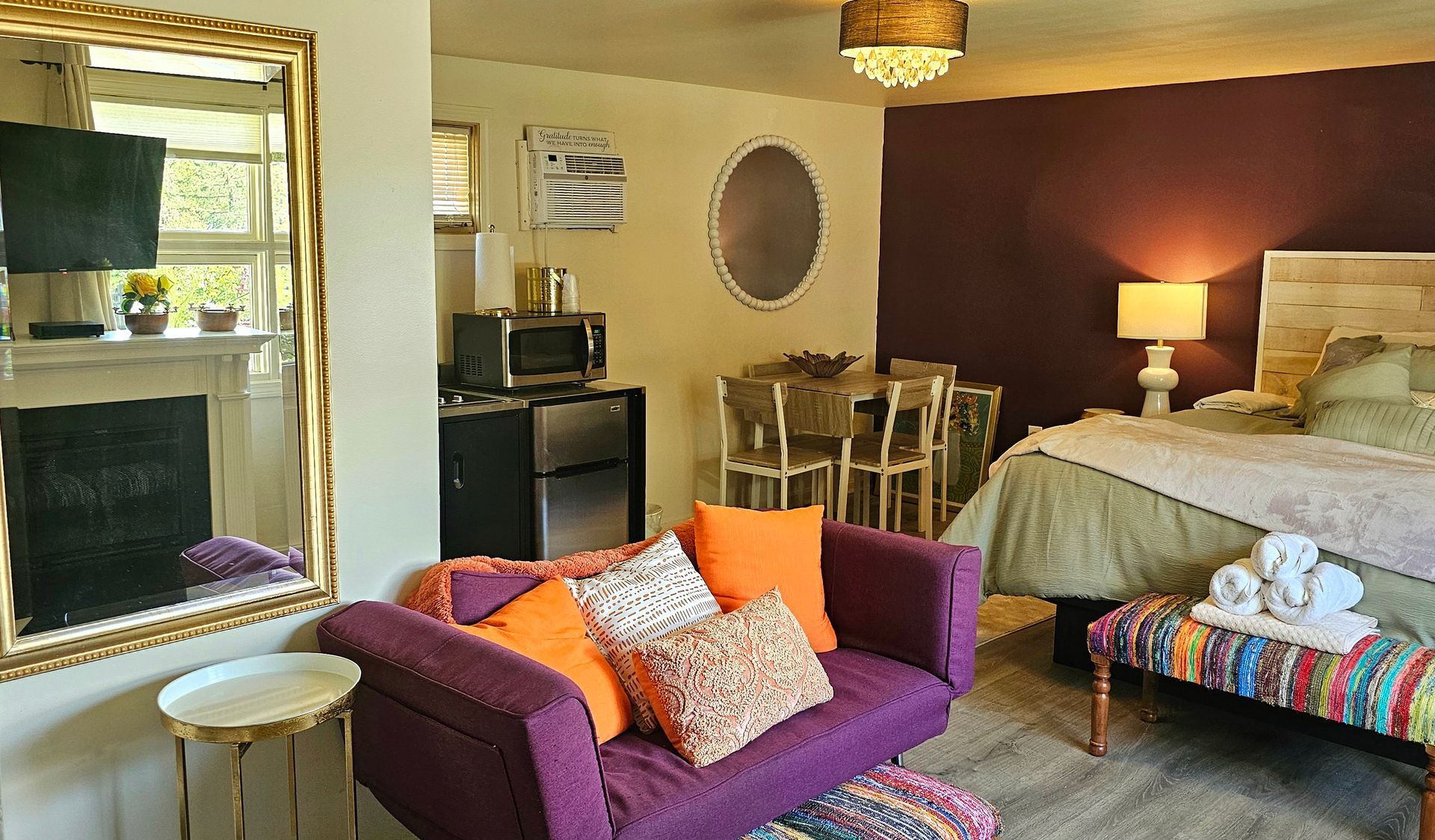 Purple couch and bed in a studio apartment.  Small kitchen, dining area, and large mirror.