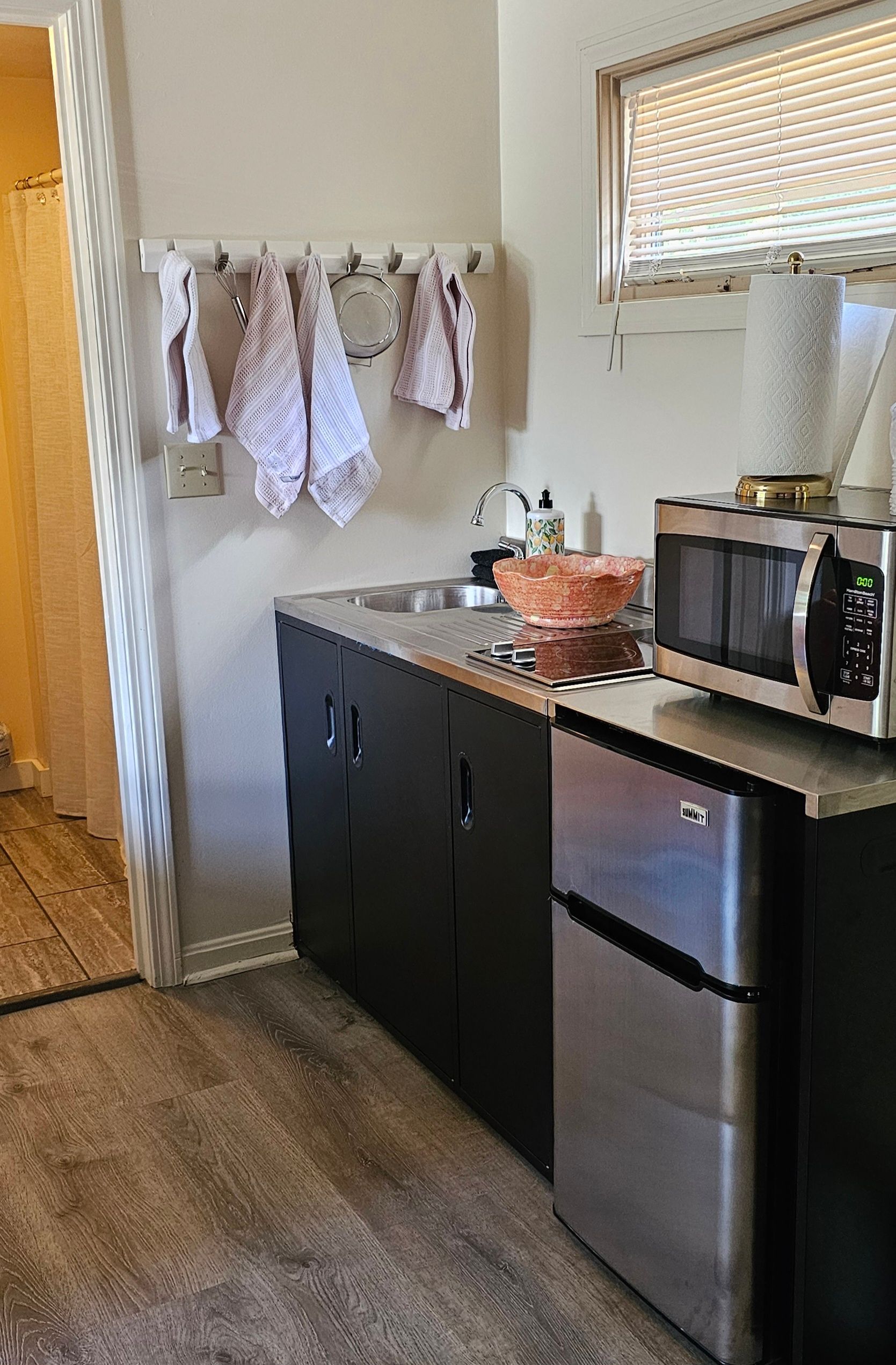 Small kitchen with dark cabinets, stainless steel mini-fridge, microwave, and sink. Dish towels hang on a wall rack.