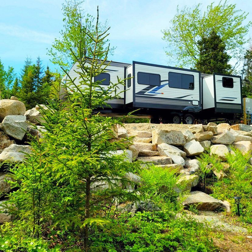 RV parked on rocky terrain, with trees and greenery under a bright, sunny sky.