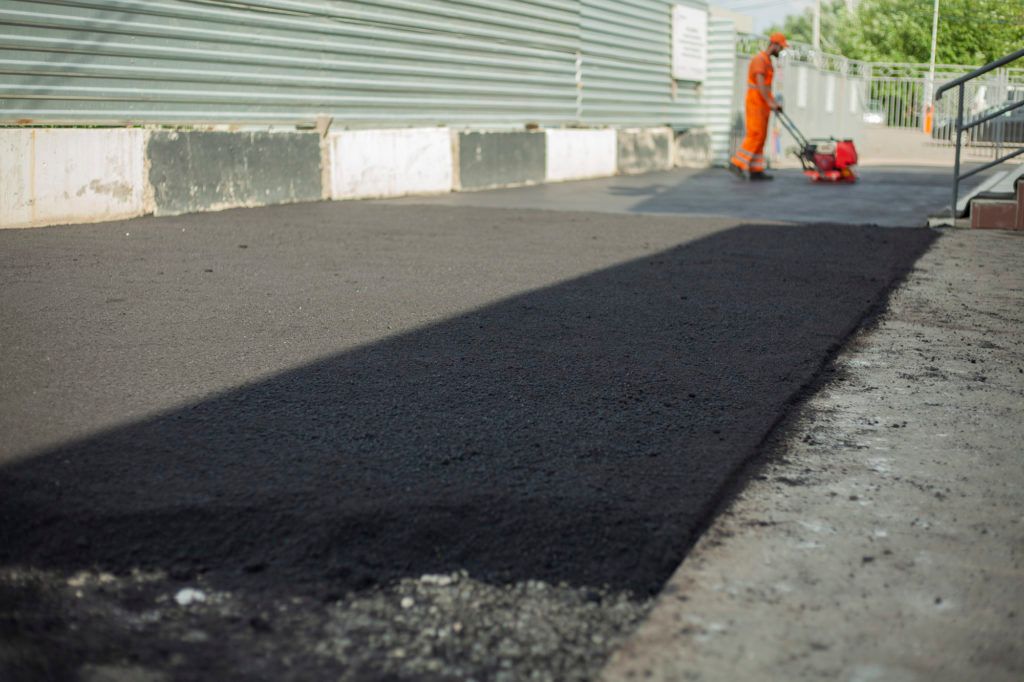 A man is laying asphalt on a sidewalk next to a building.