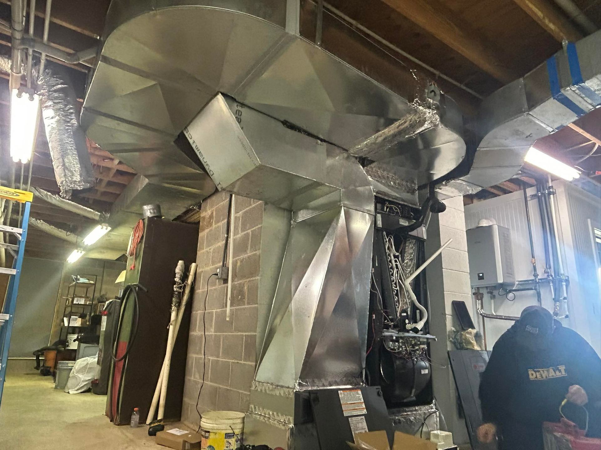 A worker installs custom sheet metal ductwork connecting to a heating system in an unfinished basement or industrial space.