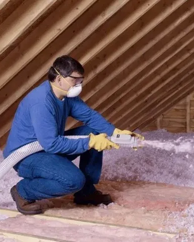 A person wearing safety gear, respirator, and gloves uses a blower hose to install loose-fill insulation in an attic.