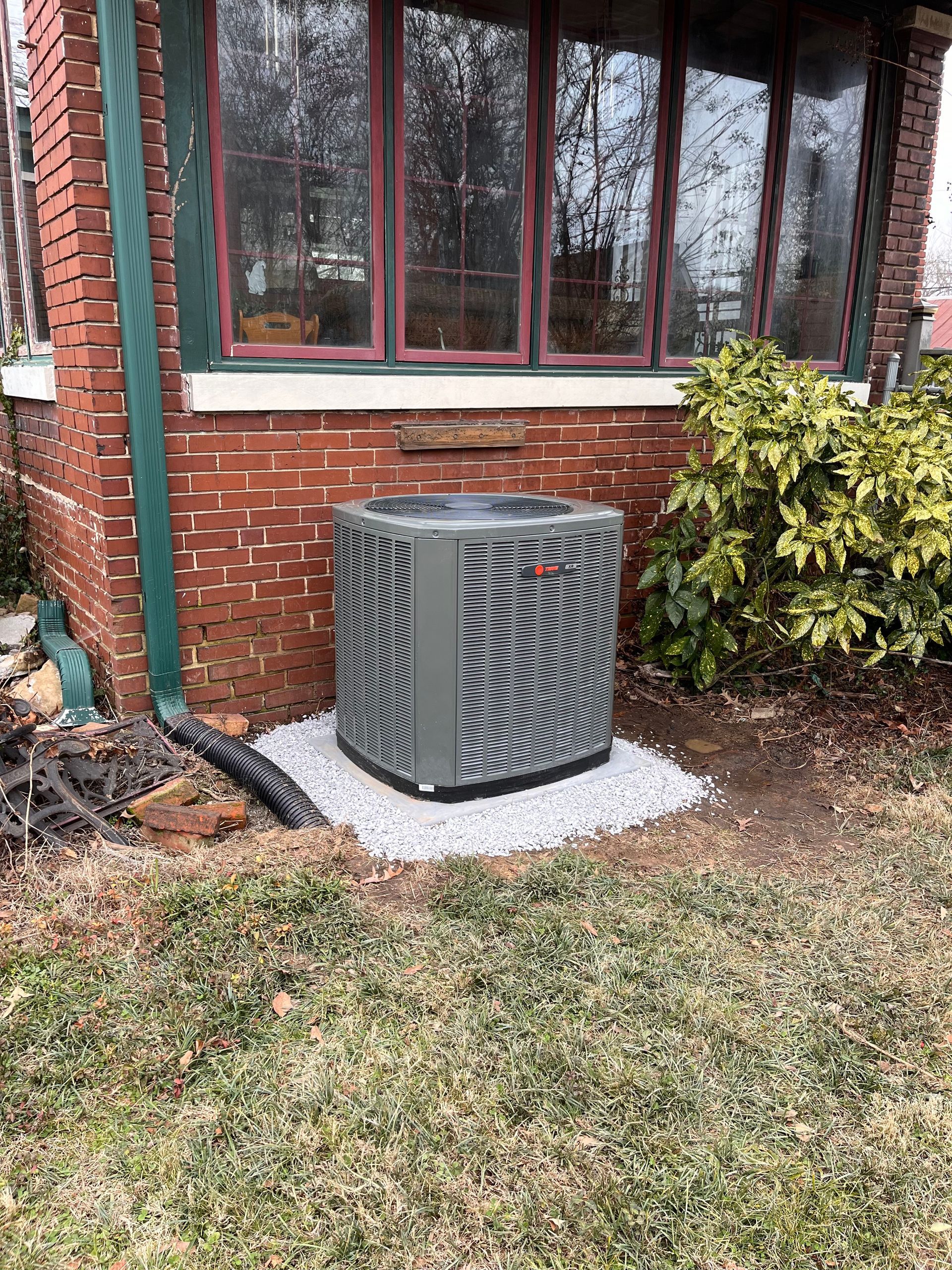 A grey air conditioning unit sits on a white gravel pad next to a red brick wall and house windows.