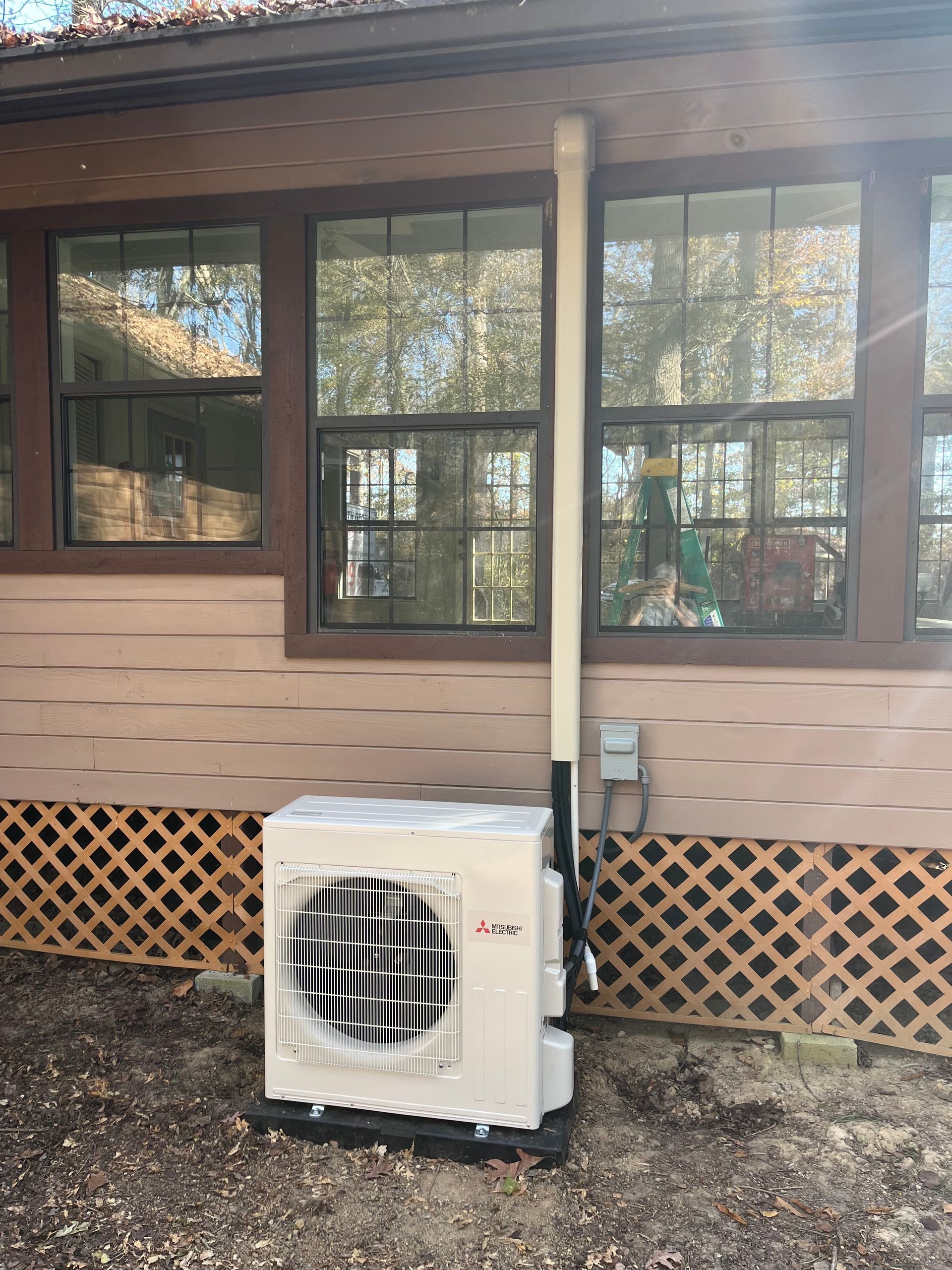 A beige mini-split heat pump unit stands on a gravel surface against a house exterior with wood lattice skirting.