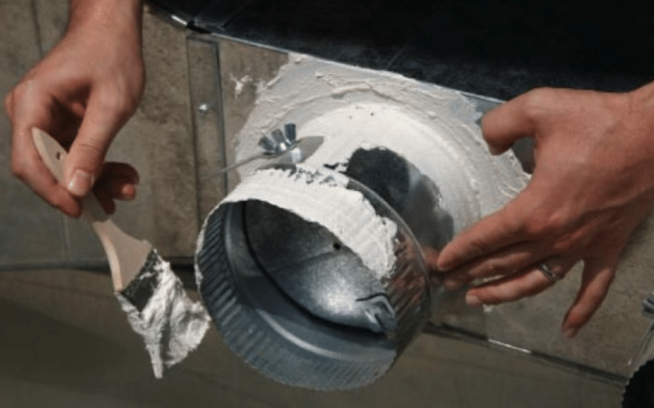A person using a brush to apply white mastic sealant around the base of a metal duct collar attached to a sheet metal box.