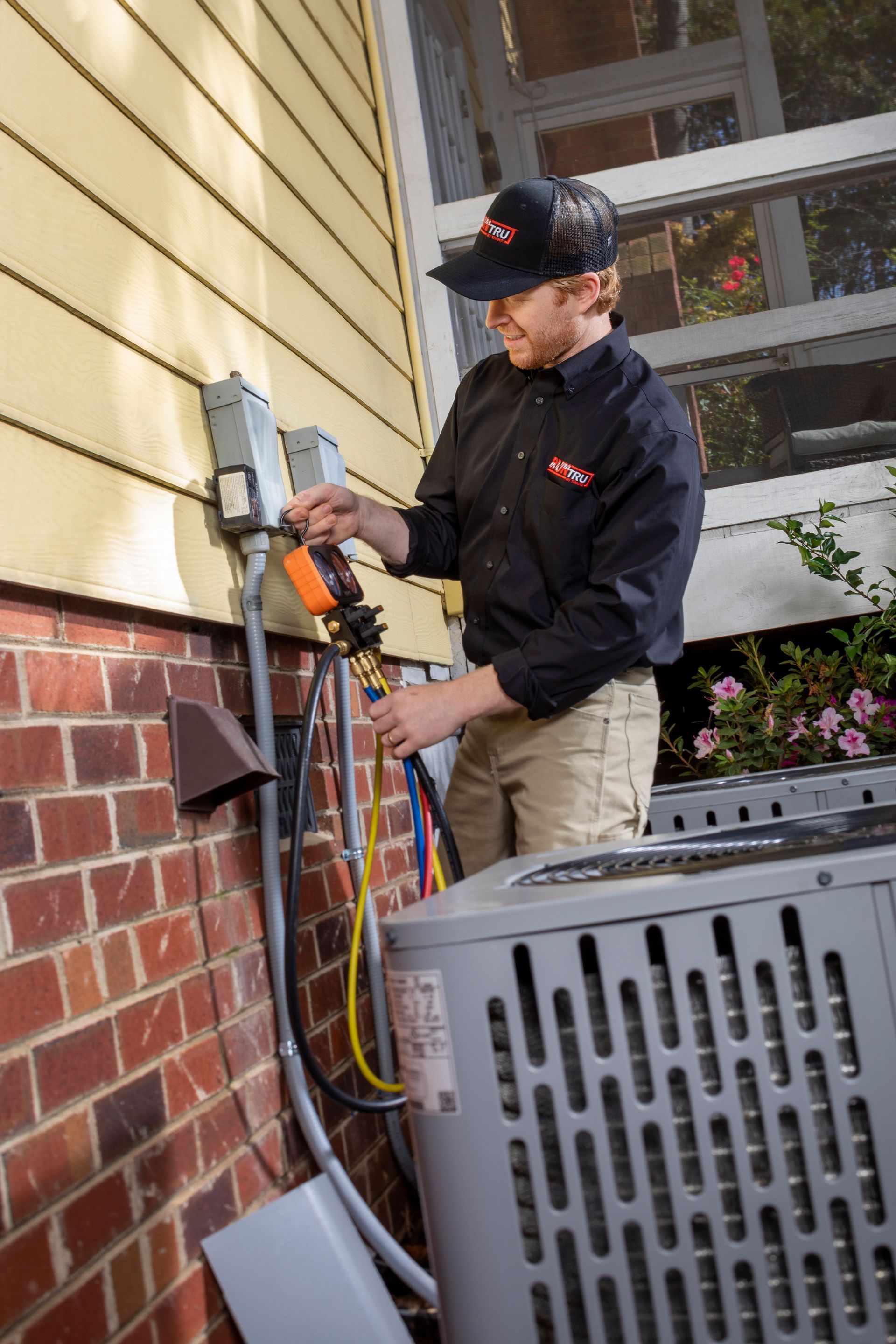 A technician in a black uniform tests the electrical disconnect box of an outdoor HVAC unit on a brick and siding house.