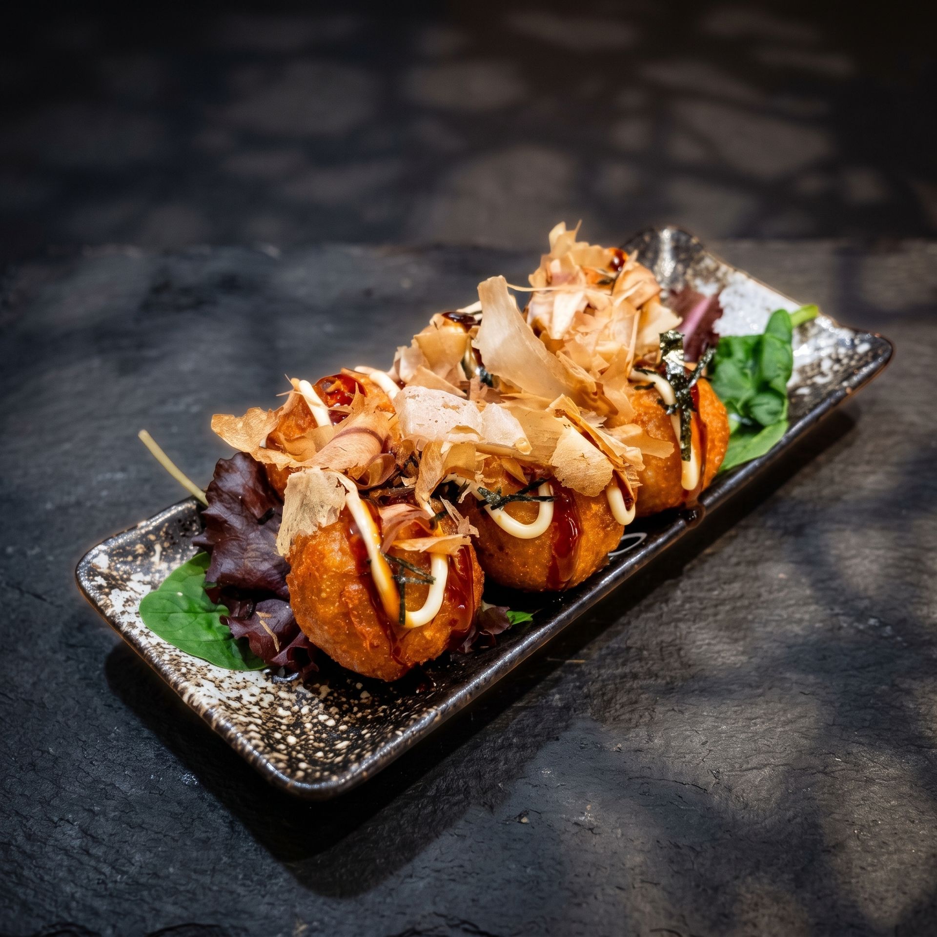 Gourmet bite-sized appetizers topped with crispy garnish on a black platter with greens, on a dark table