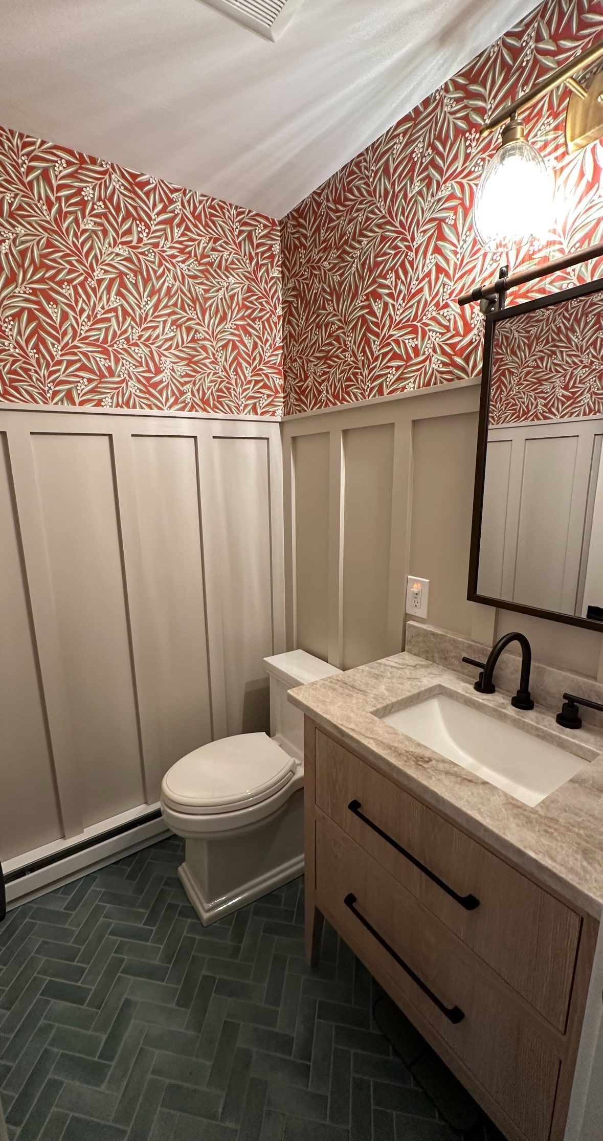 Bathroom with red patterned wallpaper, white wainscoting, and a wooden vanity on blue herringbone tile.