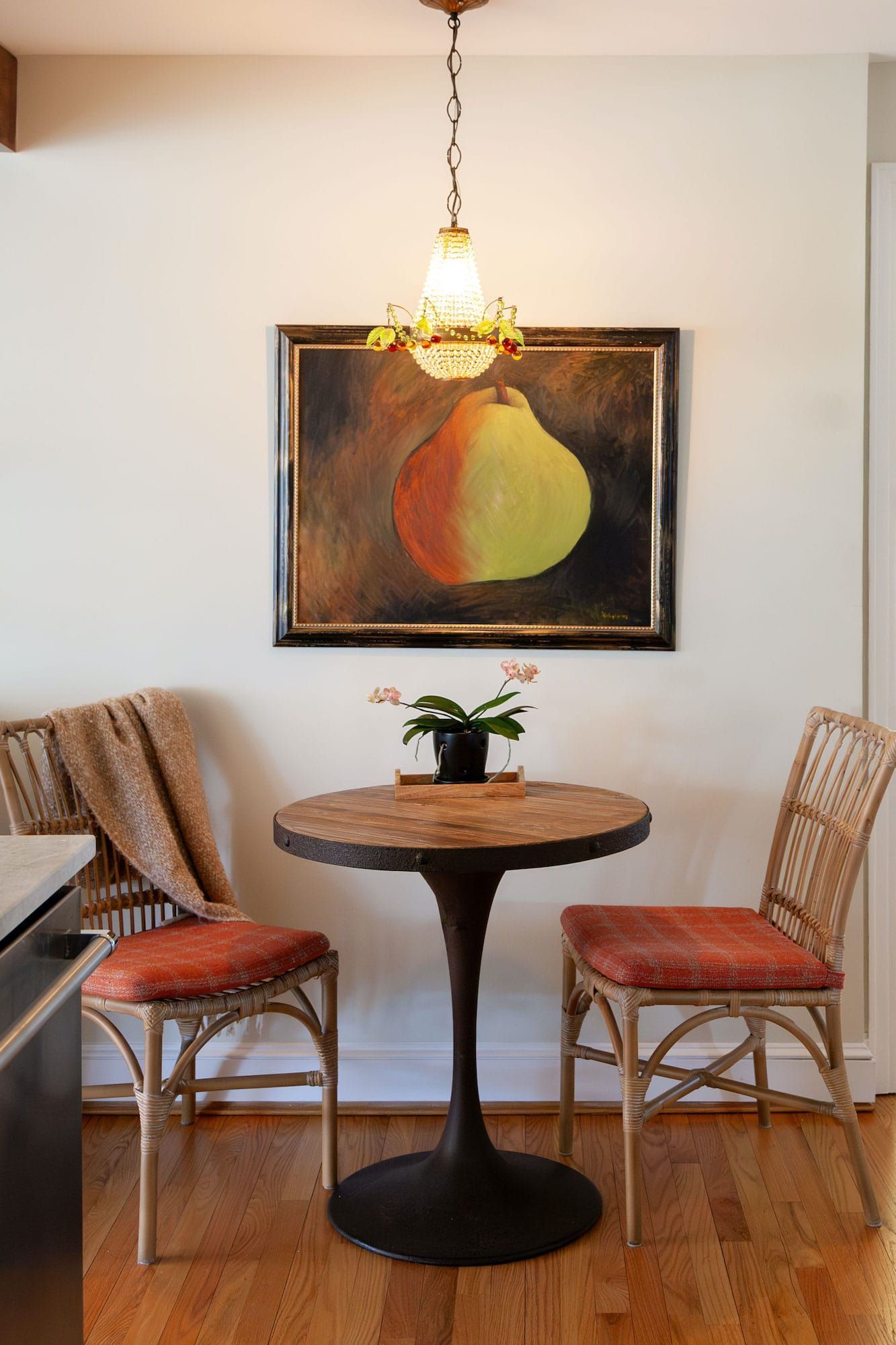 Dining nook: round table, two chairs with orange cushions, pear artwork, hanging light, wooden floor.