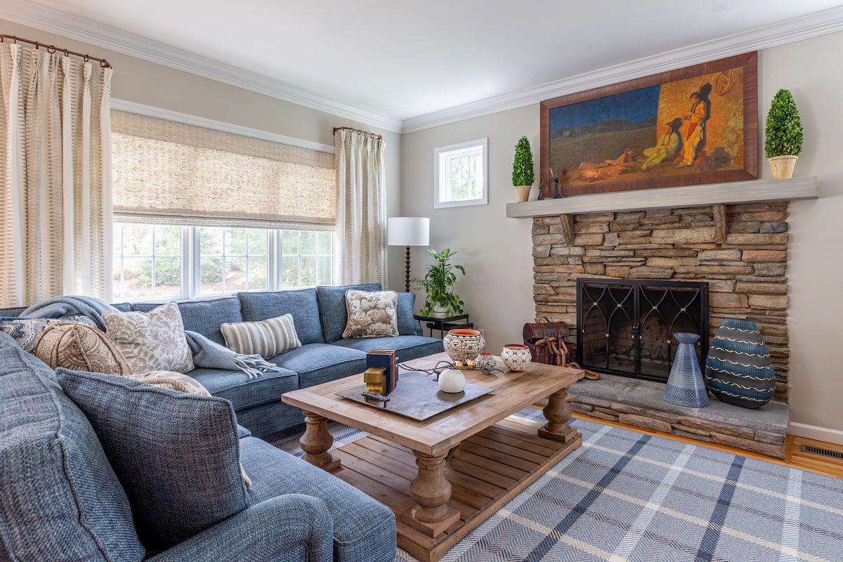 Cozy living room with blue sectional, stone fireplace, wooden coffee table, and window with beige curtains.