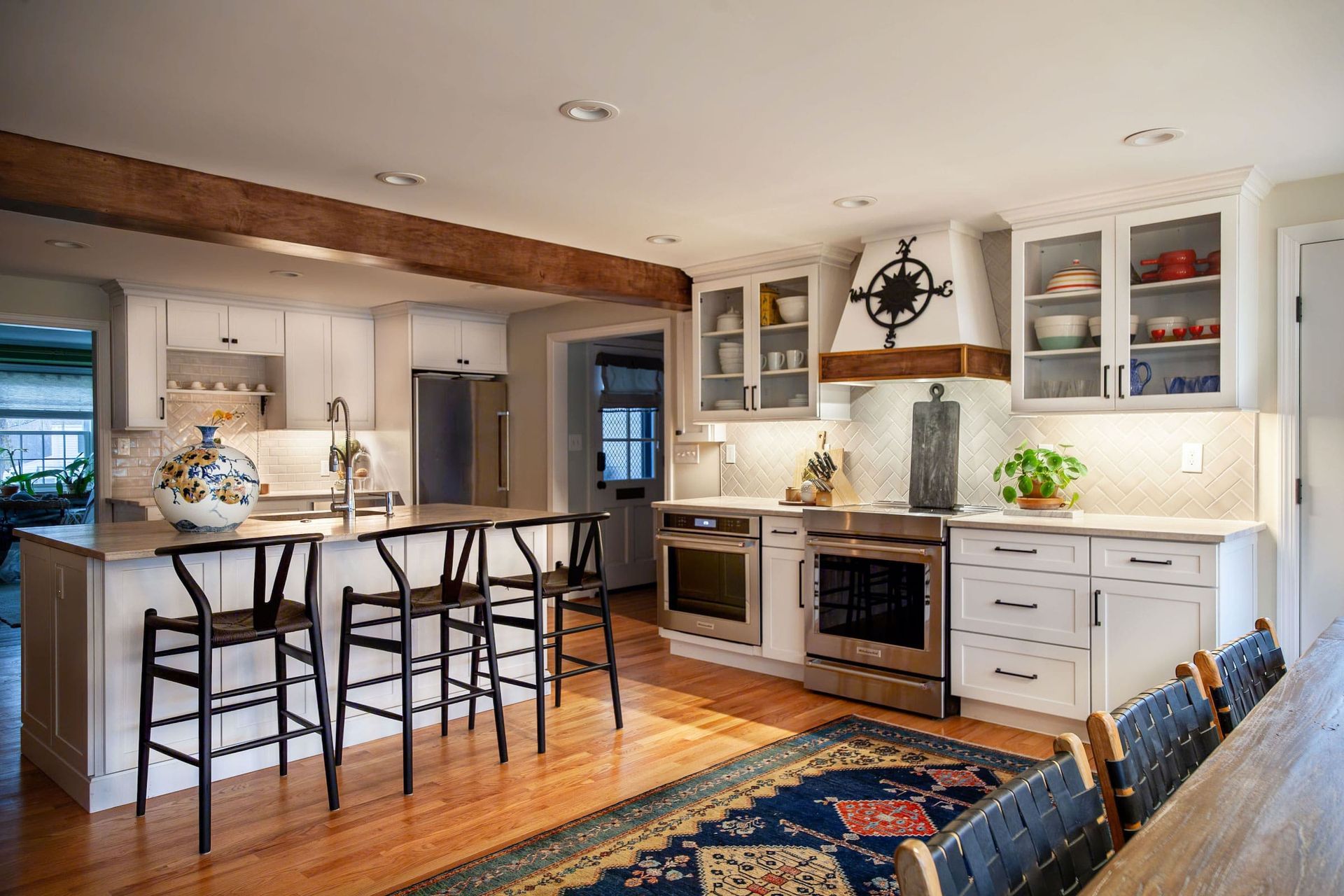 Spacious white kitchen with wooden beams, stainless steel appliances, island with black chairs, and a decorative rug.