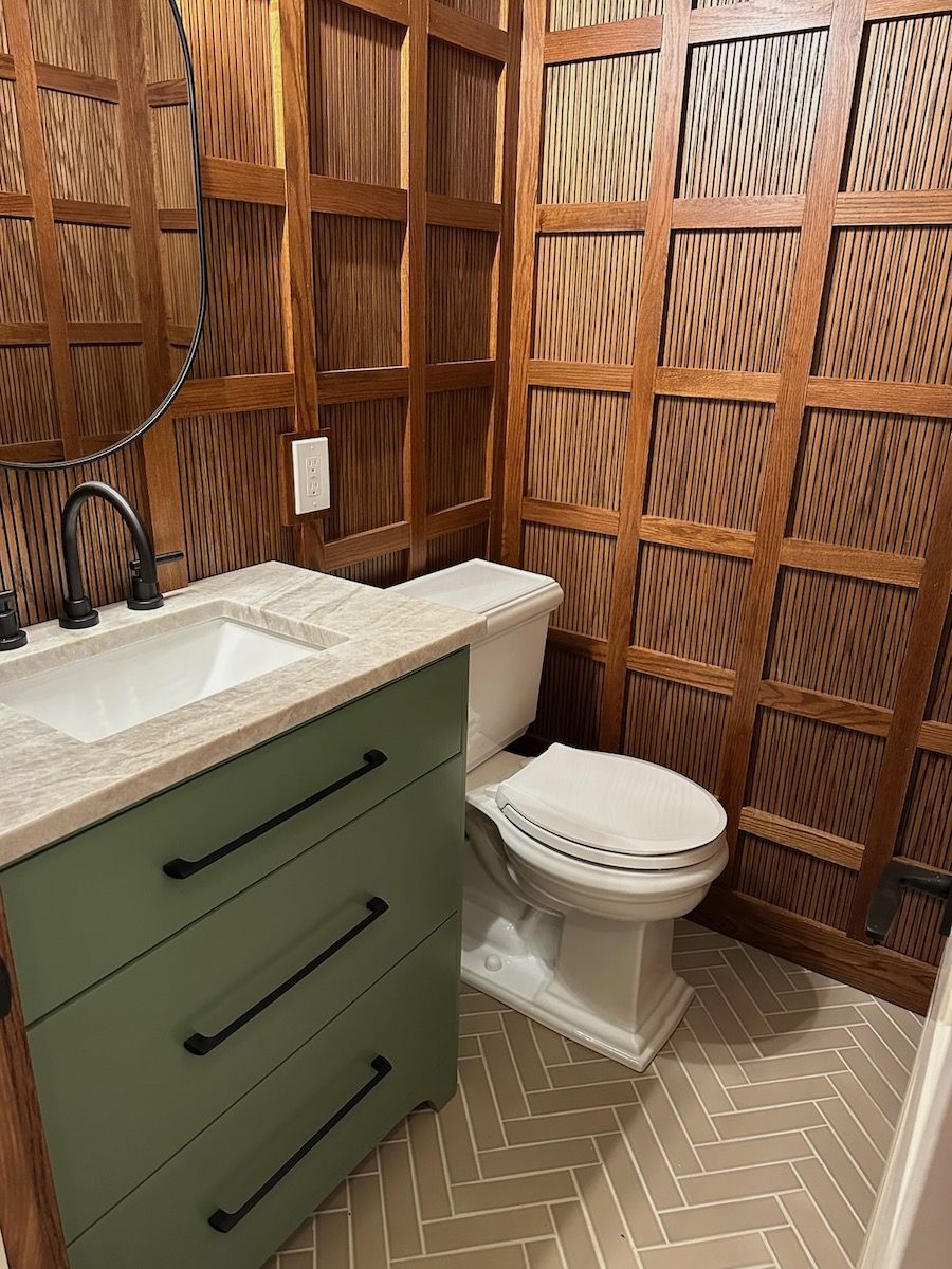 Green vanity and white toilet in a small bathroom with wood paneling and herringbone floor.