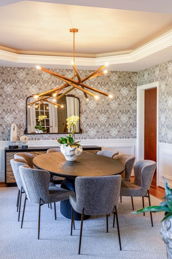 Elegant dining room with a large round table, chairs, and gold chandelier. Patterned wallpaper and a decorative sideboard are present.