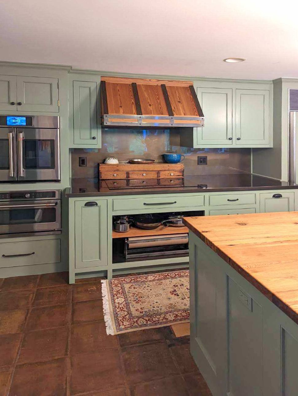A rustic kitchen with green cabinets, wooden accents, and a rug on a tiled floor.