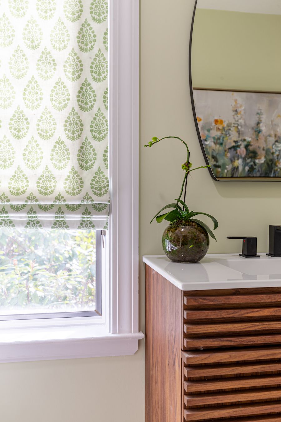 Bathroom with patterned window shade, wooden cabinet, mirror, and plant.