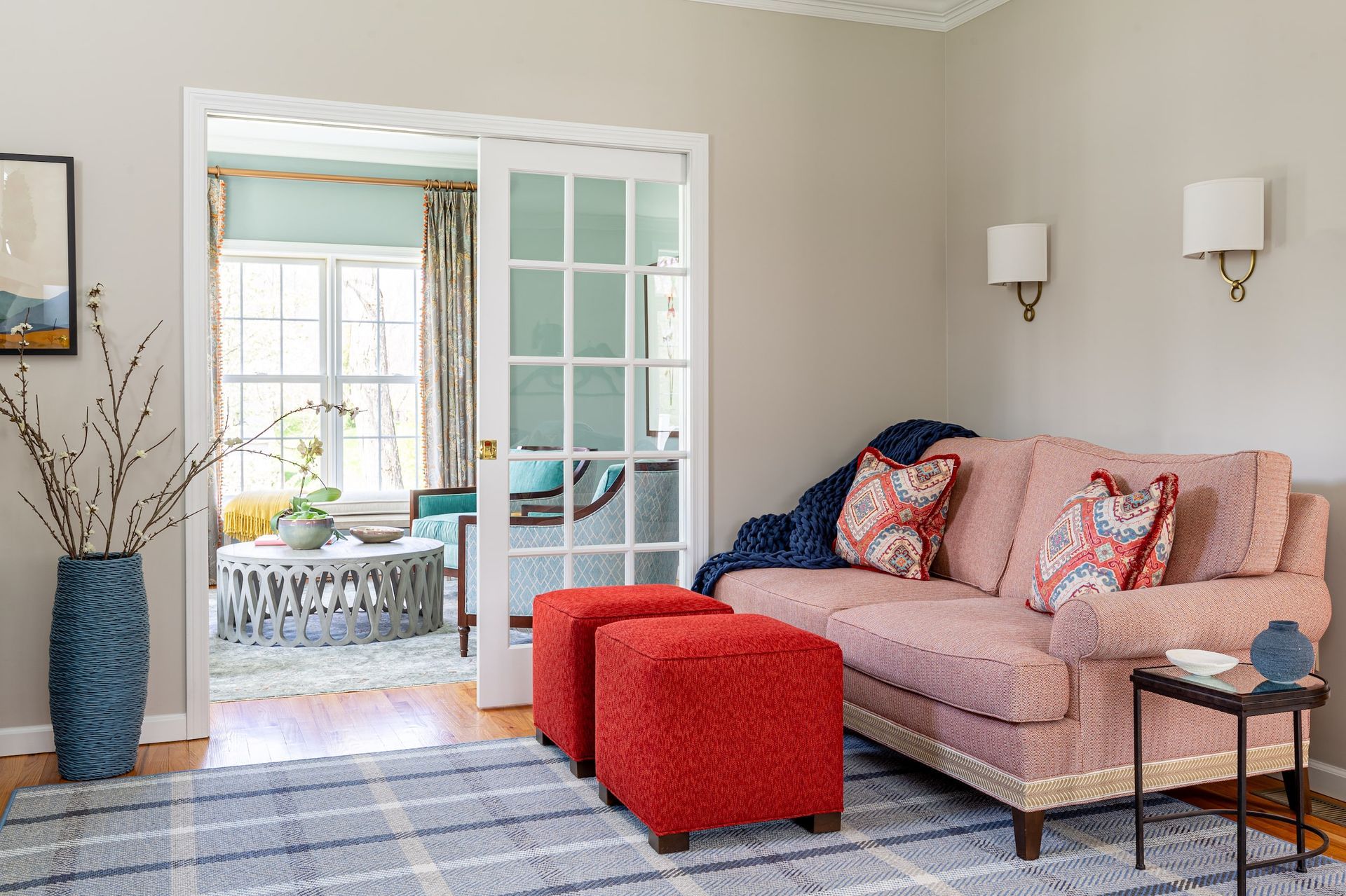 Cozy living room: pink sofa, red ottomans, blue rug, doorway to another room with windows, beige walls.