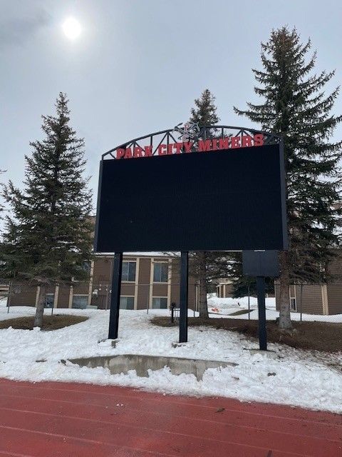 Black scoreboard in front of a building with snowy ground and a running track. 
