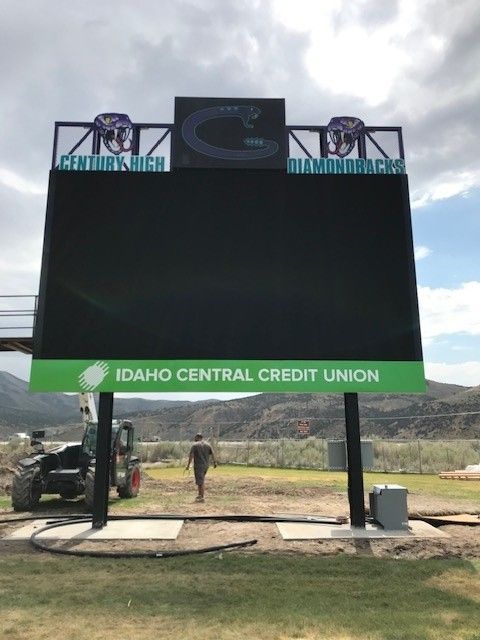 Large digital scoreboard at Centennial High School, with a person walking by. Green Idaho Central Credit Union sign below the black screen.