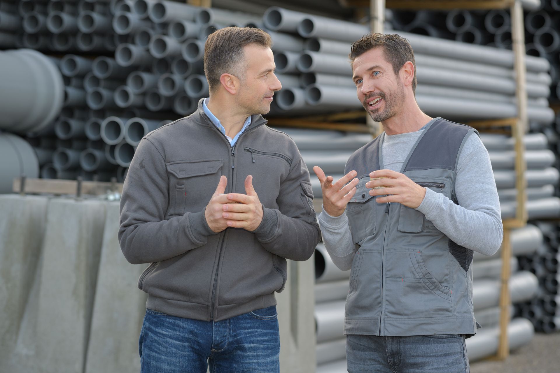 Two plumbing professionals discussing a project while standing near stacked pipes at a work site. Two plumbing professionals discussing a project while standing near stacked pipes at a work site.