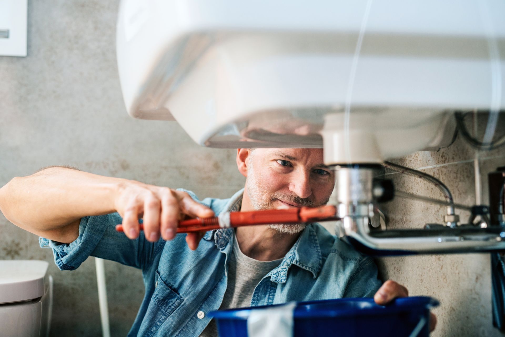 A person fixing a sink with a wrench and holding a blue bucket underneath.