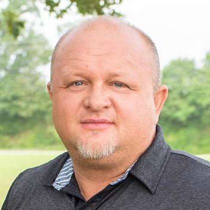 Portrait of a smiling bald man in a dark shirt, outdoors with greenery in the background