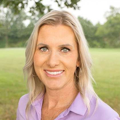 Smiling woman with blonde hair in a purple shirt standing outdoors in a green park