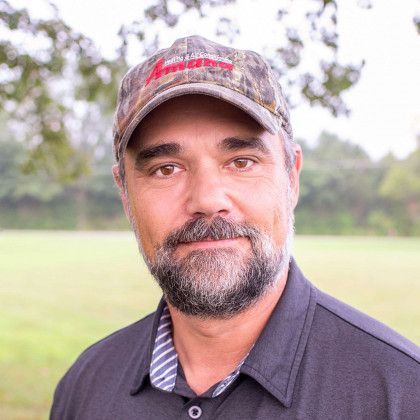 Portrait of a smiling man in a camouflage cap outdoors, with a grassy field and trees behind him