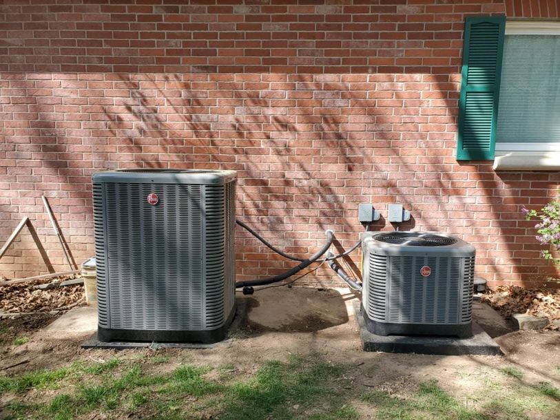 Two outdoor air conditioning units beside a brick wall, with a green shutter window above.