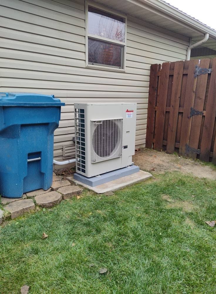 Outdoor air conditioner unit beside a beige house, next to a blue trash bin and wooden fence.