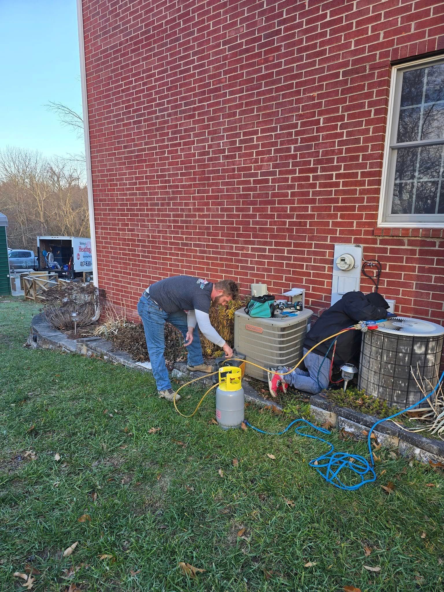 Two people work with tools and hoses beside a red brick building in a grassy yard.