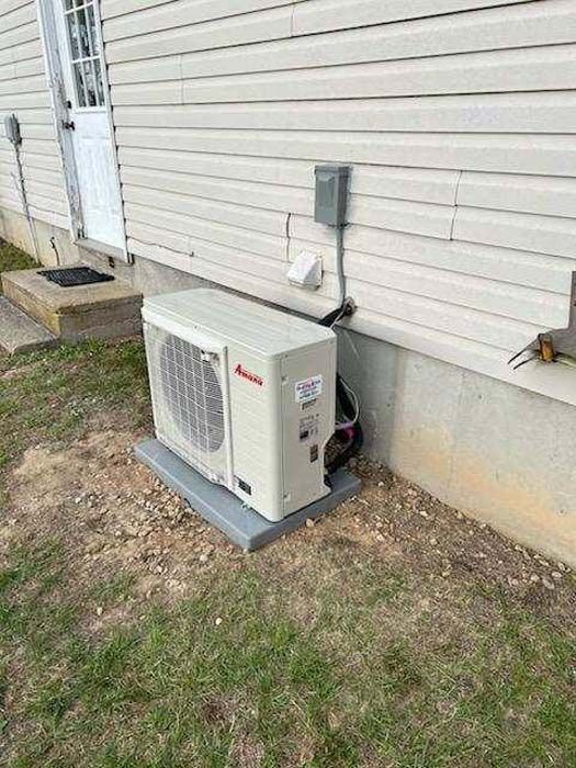 Outdoor heat pump unit beside a house wall, with electrical conduit and patchy grass nearby