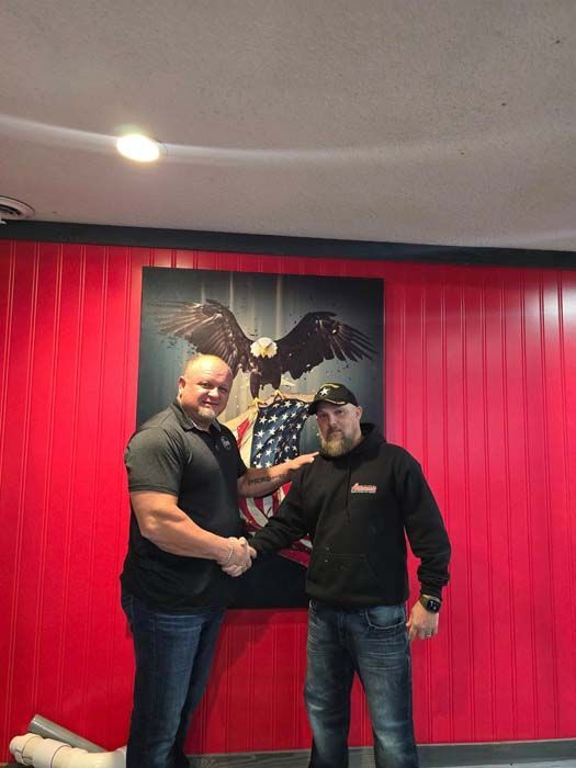 Two men shaking hands in front of a red wall and eagle artwork.