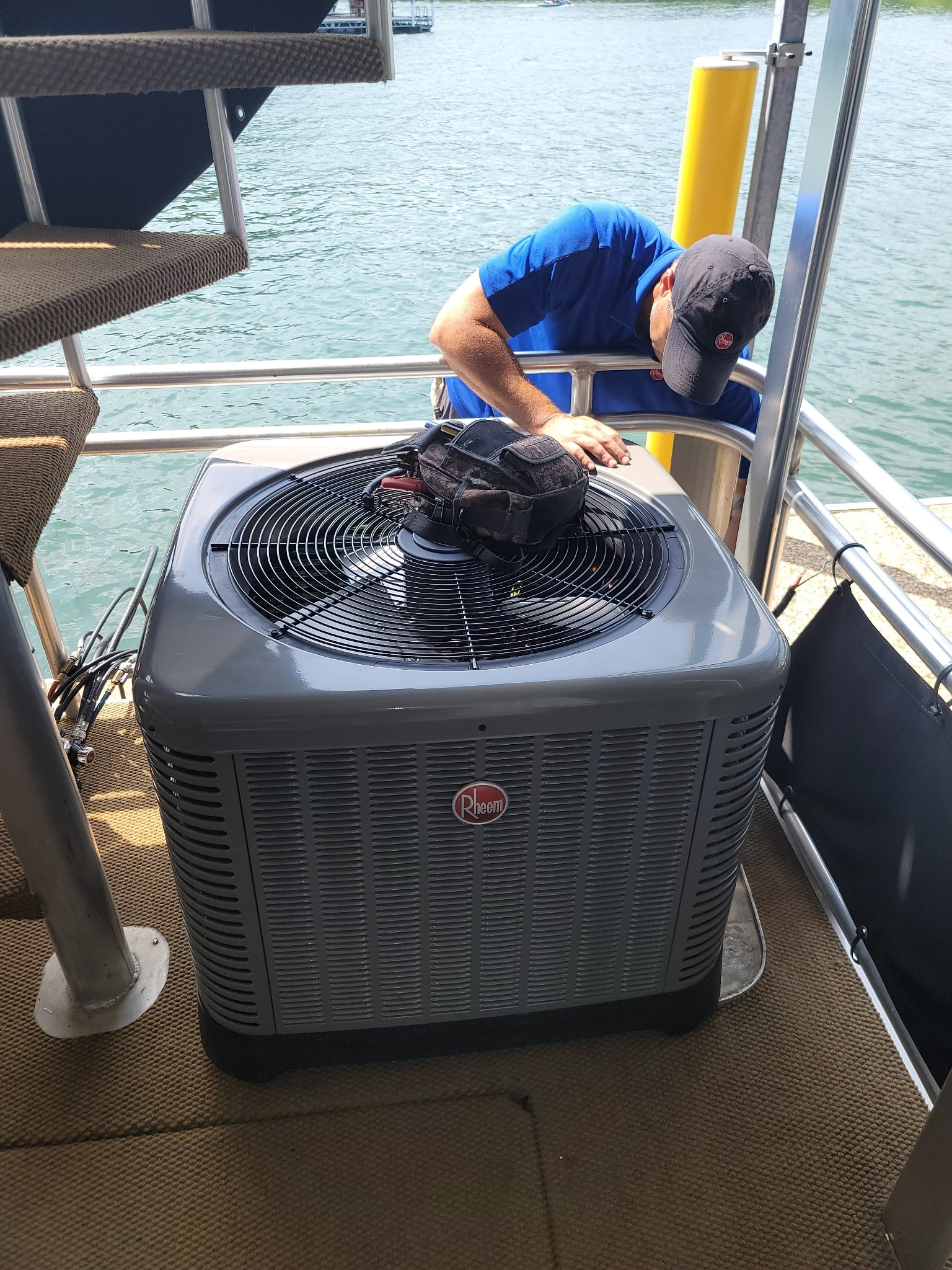 Technician servicing a rooftop air conditioner beside water, using a tool on the unit.