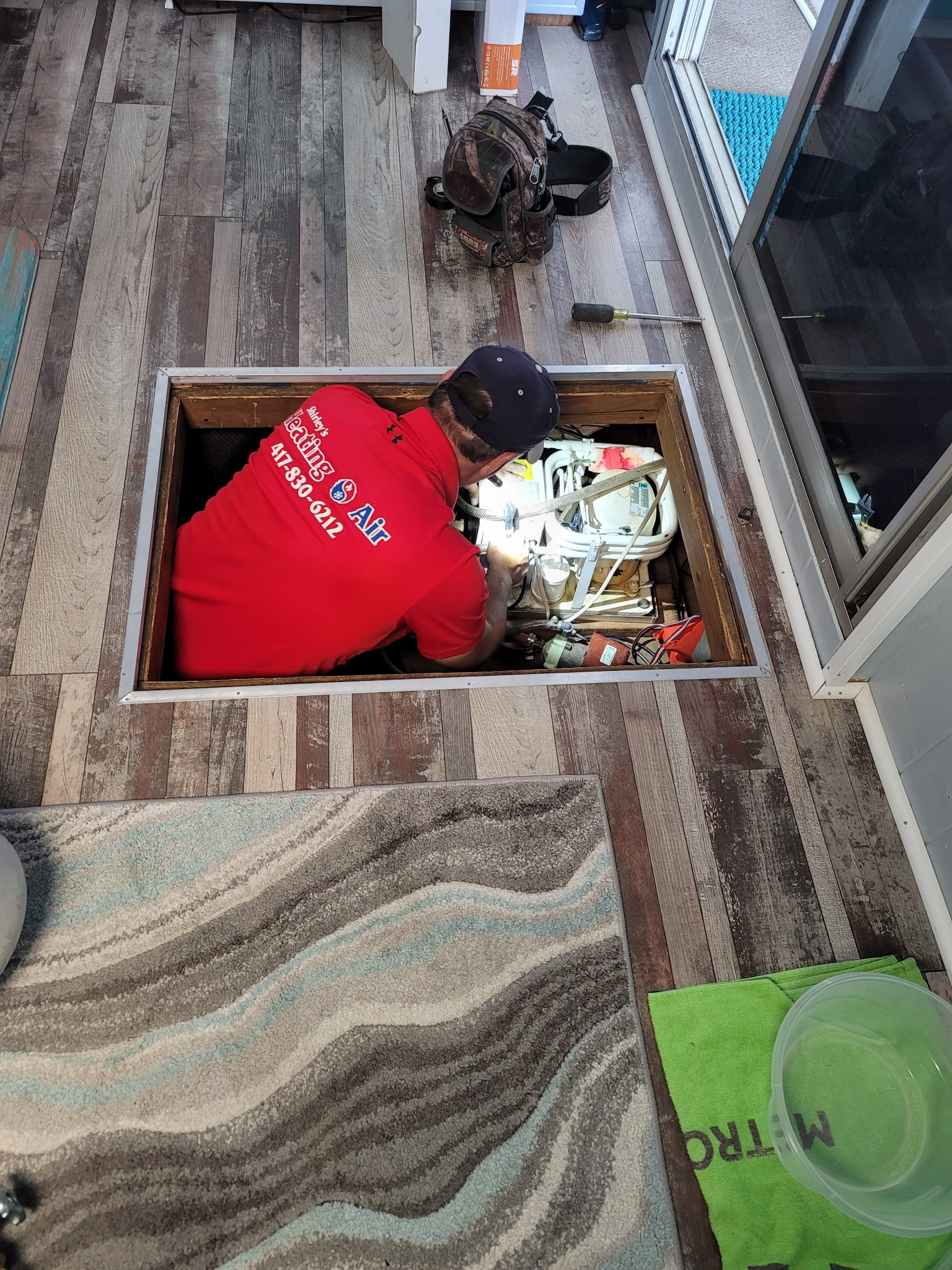Person in red shirt leaning into a floor hatch with cables and tools in a room near a sliding glass door.