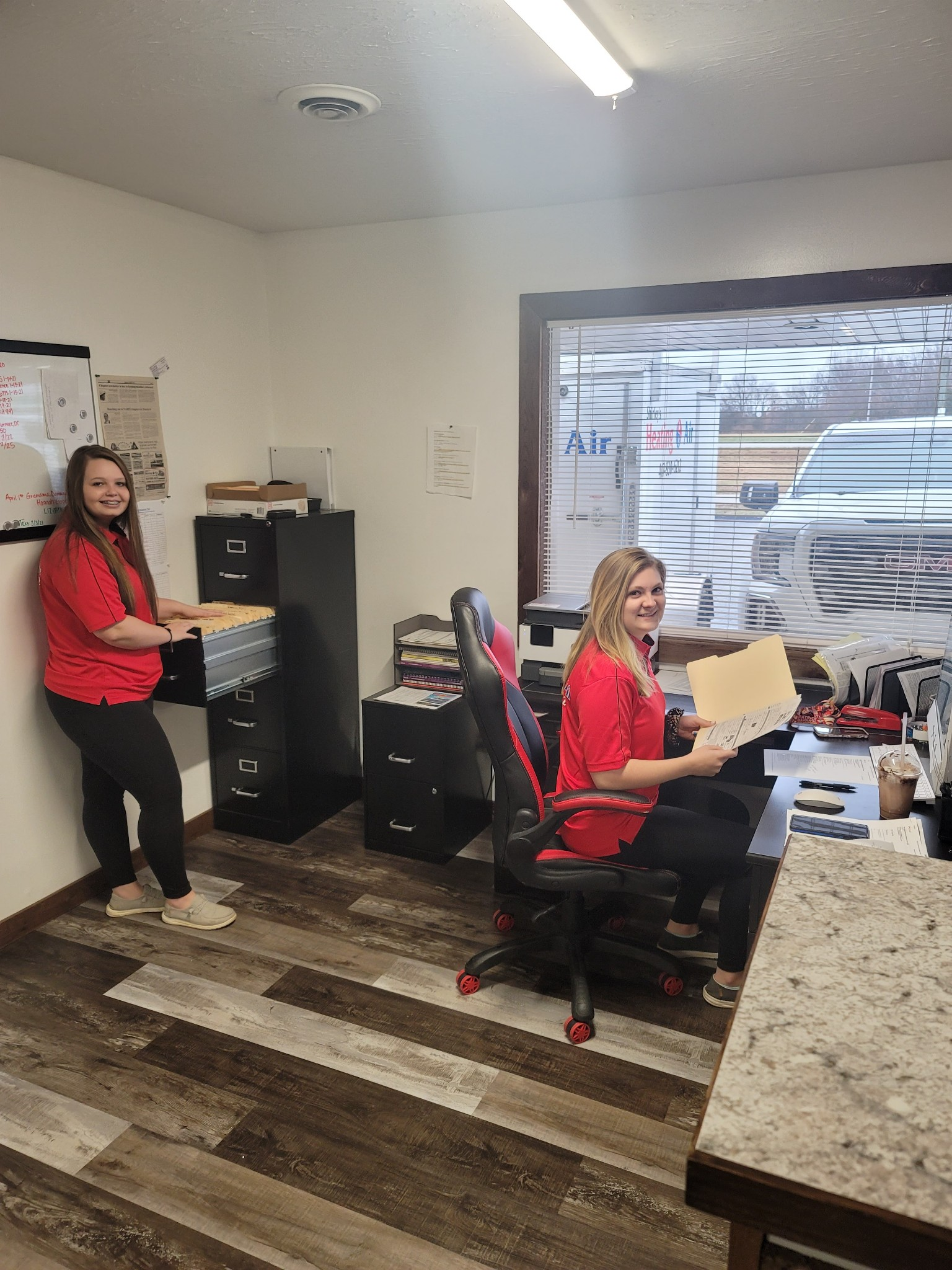 Two people in a small office, one seated at a desk and one standing near a file cabinet.