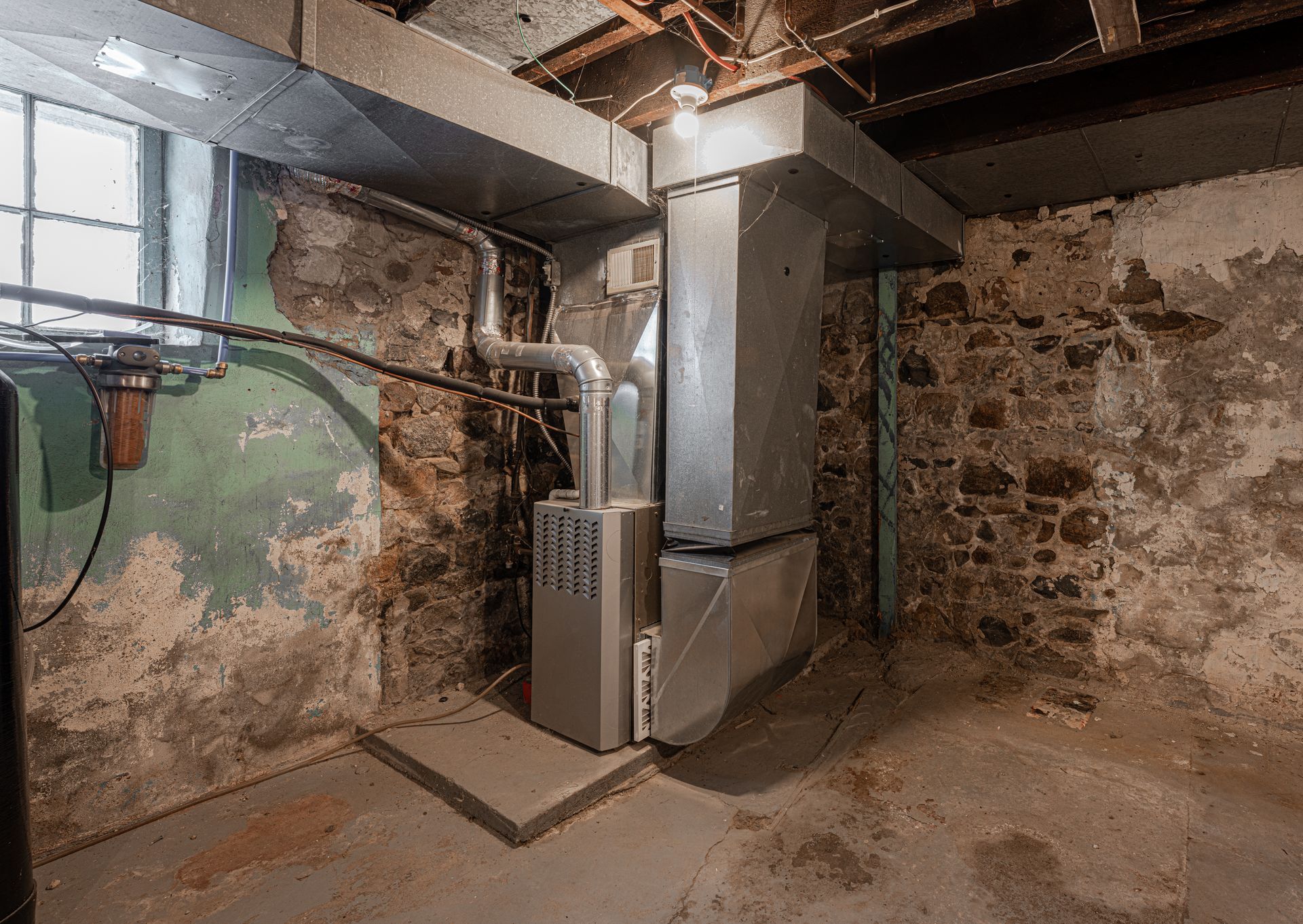 Basement furnace and ductwork beside rough stone and peeling green walls