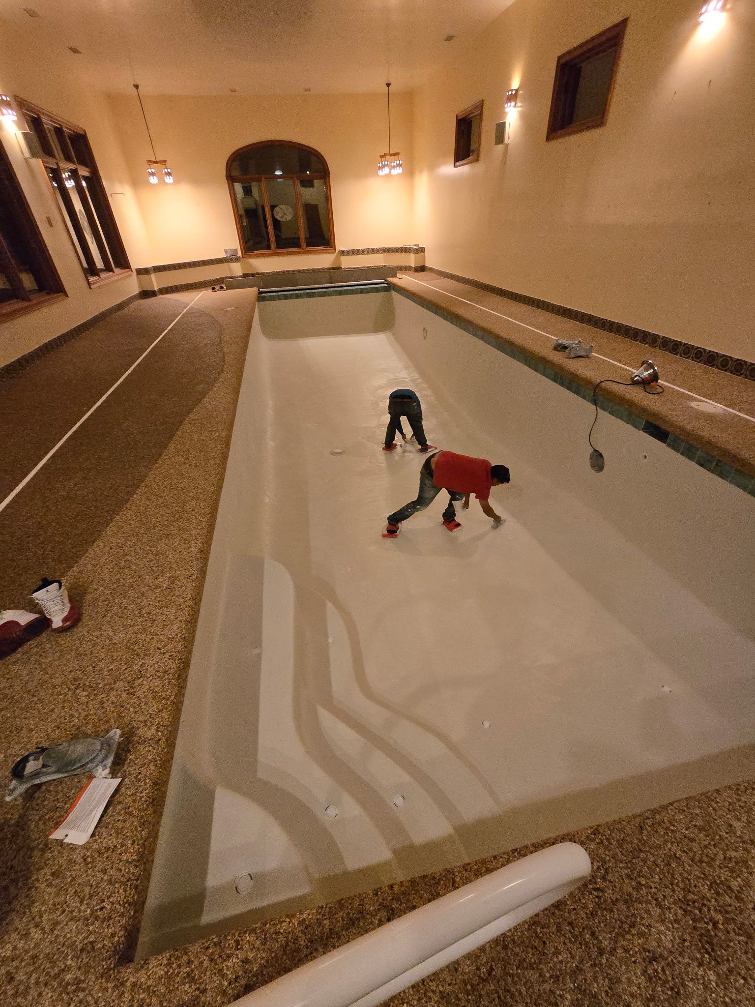 An indoor empty pool with two workers cleaning the floor, one in red and one in black.