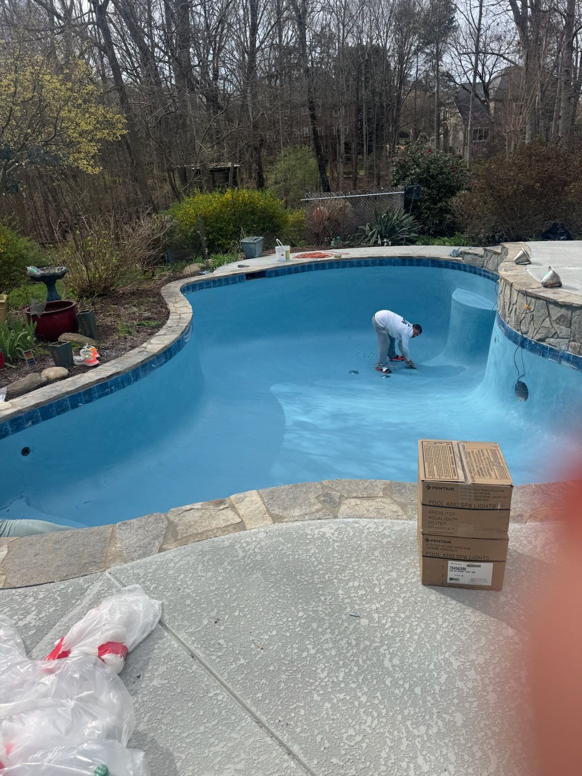 Person cleaning a blue backyard pool with a long brush, surrounded by trees and stone decking.